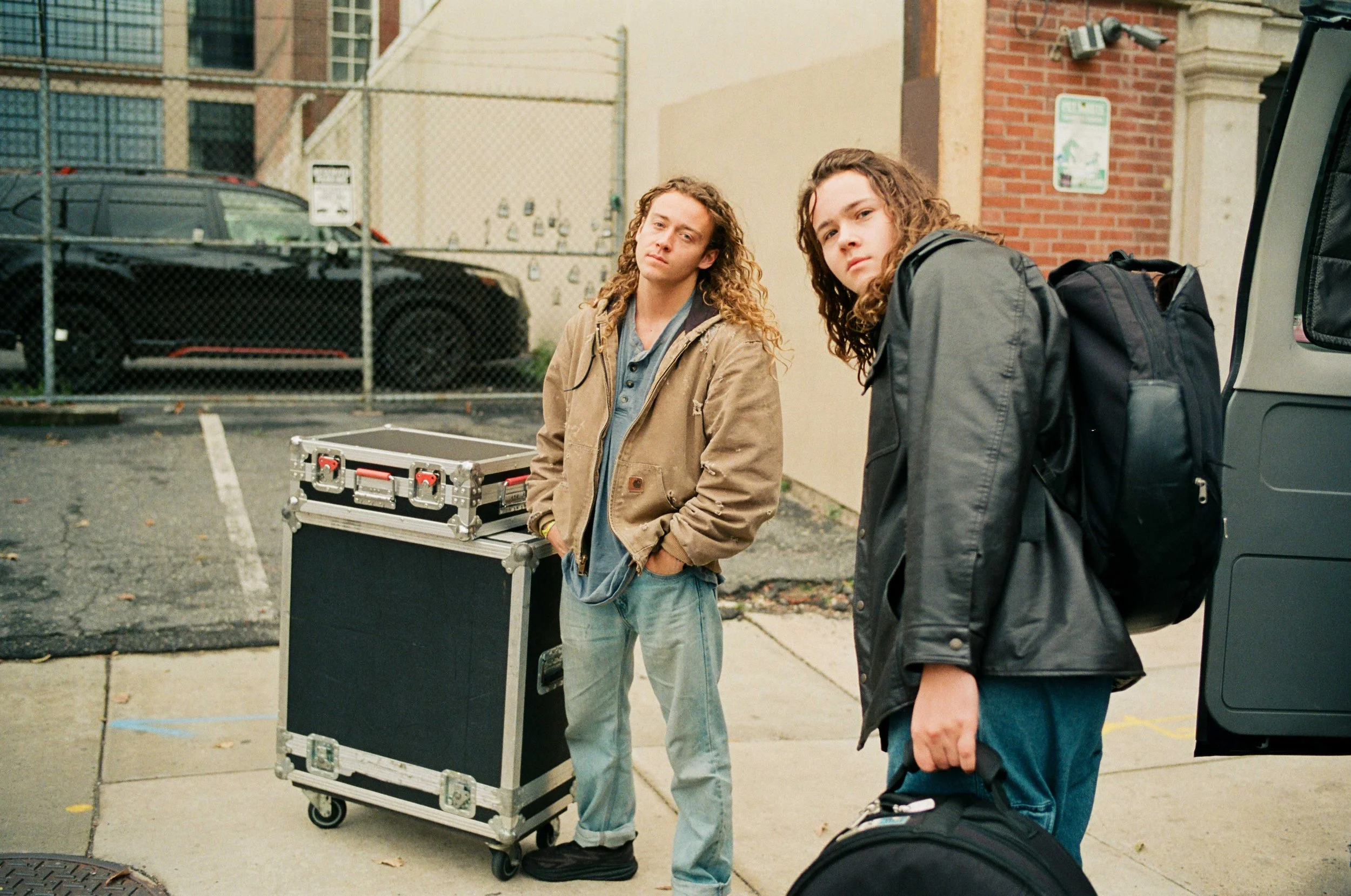 Two young men with curly hair, wearing casual jackets, standing outside in an urban area next to a vehicle and equipment case.