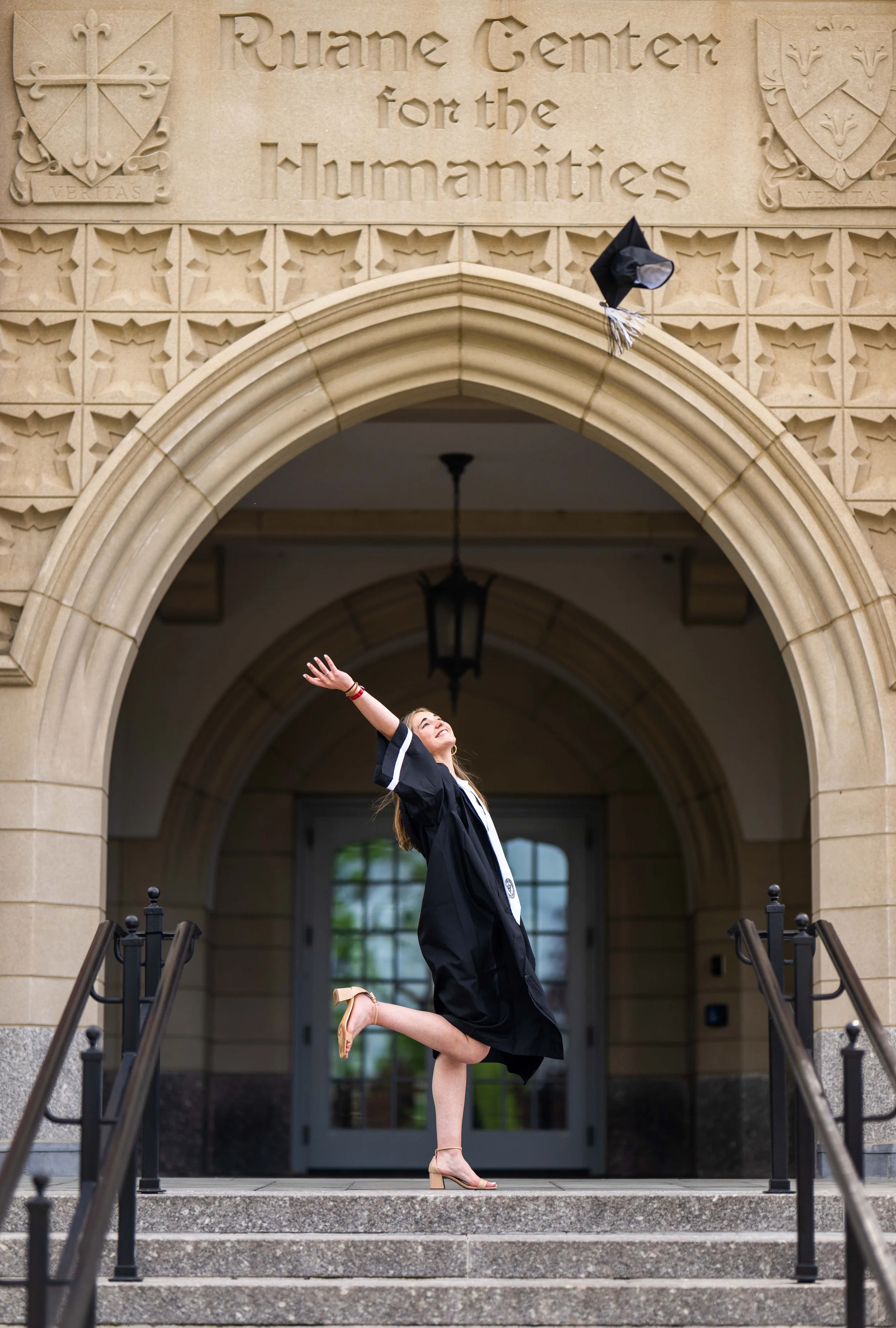 A young woman in a graduation gown and heels celebrating on the steps of the Ruane Center for the Humanities, throwing her graduation cap in the air with a joyful expression.