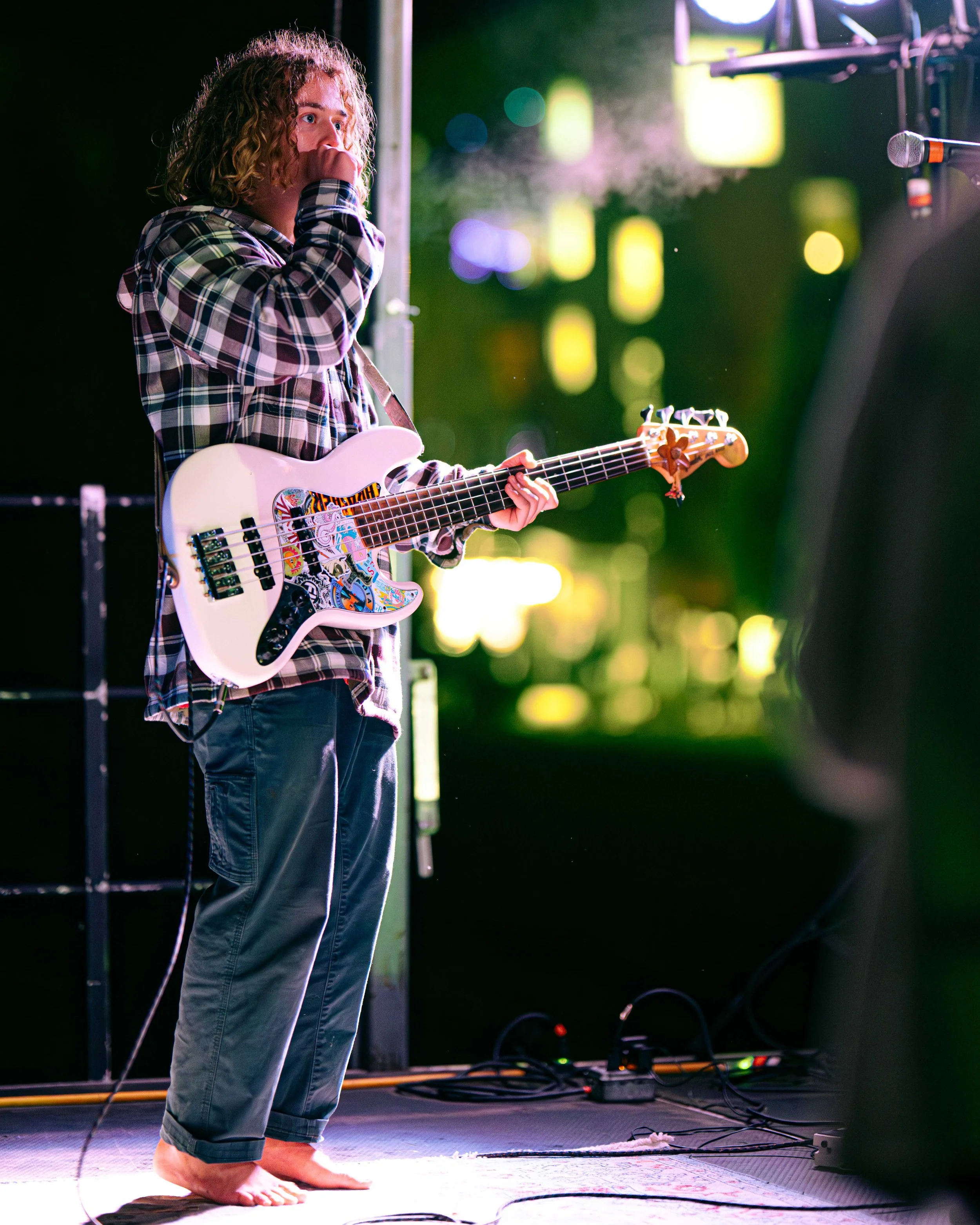 A person with curly hair wearing a plaid flannel shirt and jeans playing a white electric guitar with colorful stickers on stage at night, barefoot, with a microphone nearby and equipment on the floor.