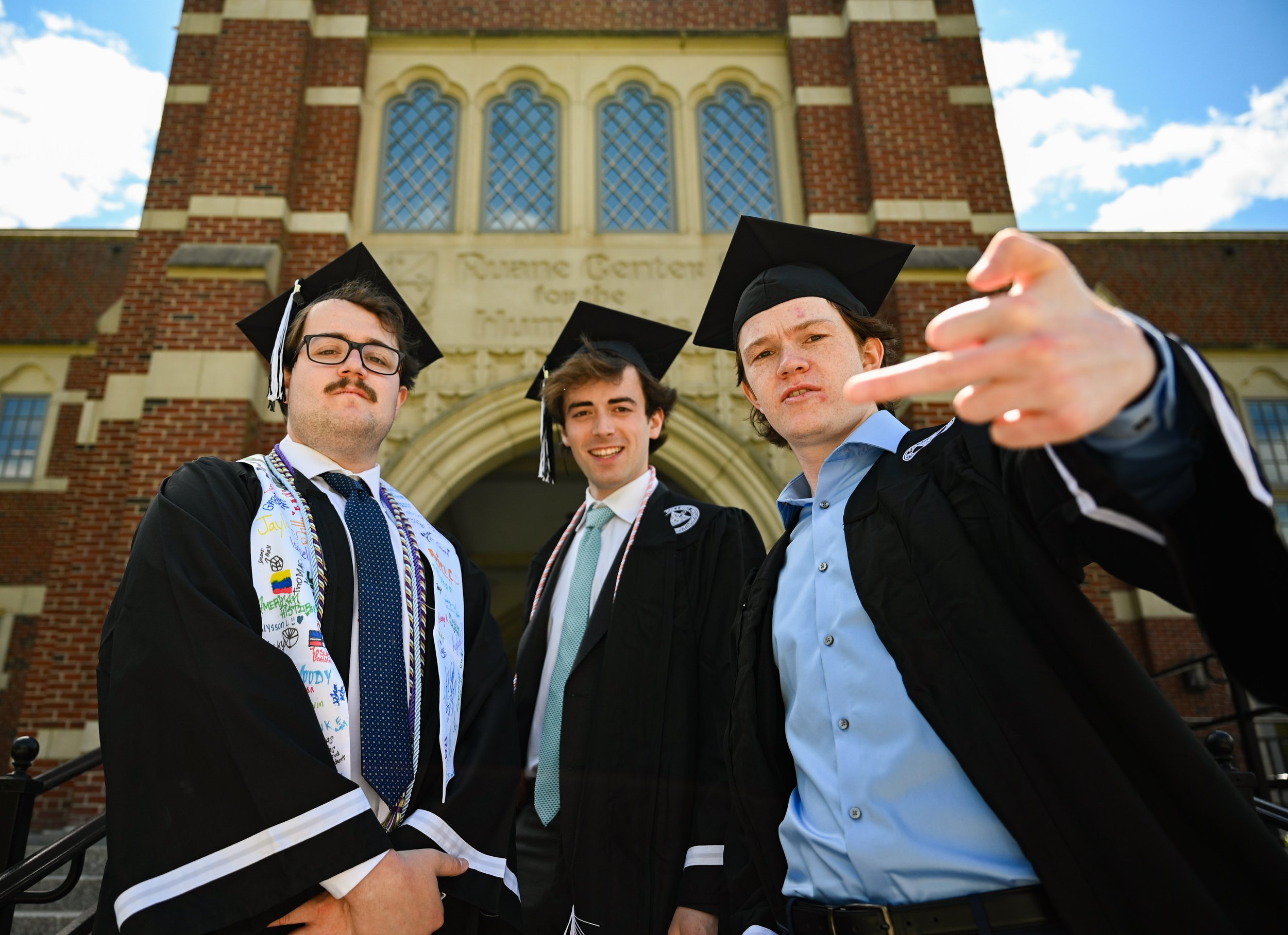 Three young men in graduation caps and gowns standing in front of a university building, smiling and making gestures at the camera.