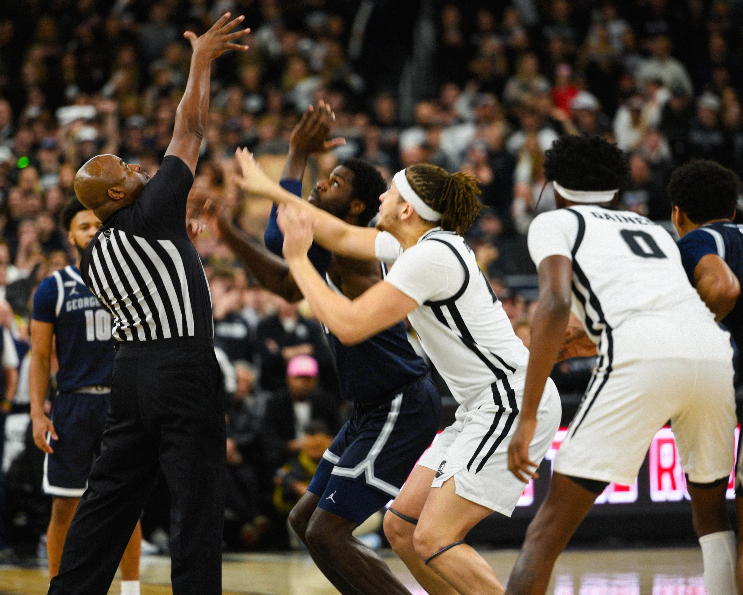 A heated basketball game with players and a referee fighting for possession of the ball on the court, with a crowded audience in the background.
