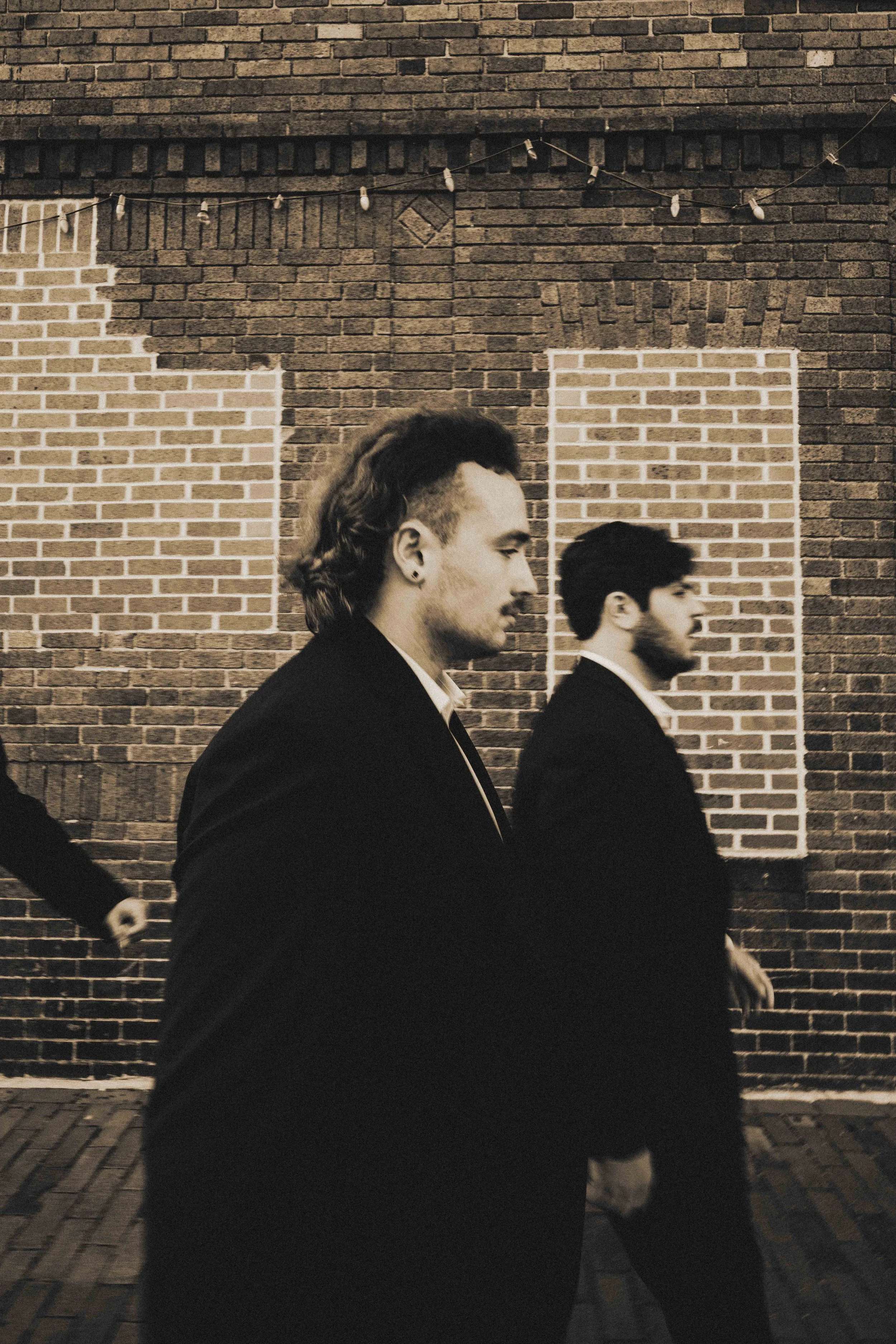 Two men in suits walking past a brick wall with a cross painted on it, in black and white.