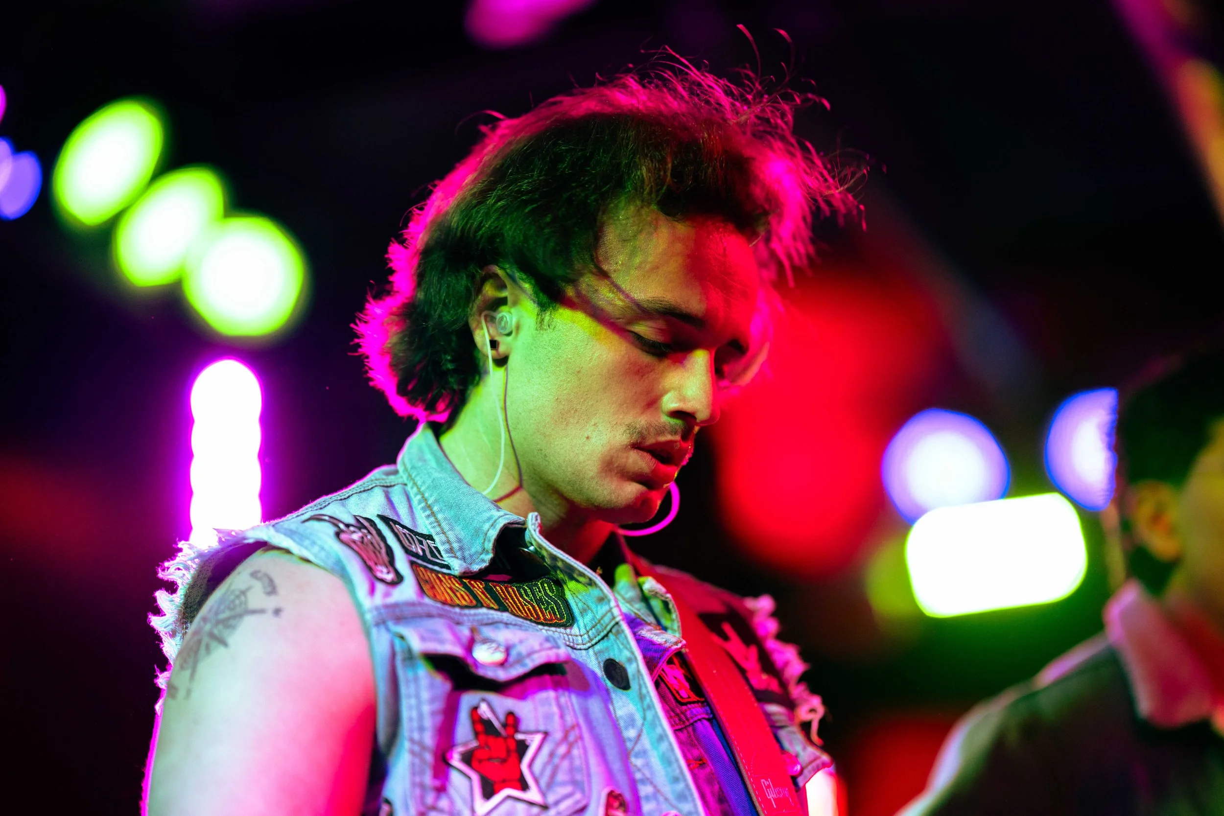 A young person with wavy hair, wearing a sleeveless denim jacket decorated with patches, inside a neon-lit environment.