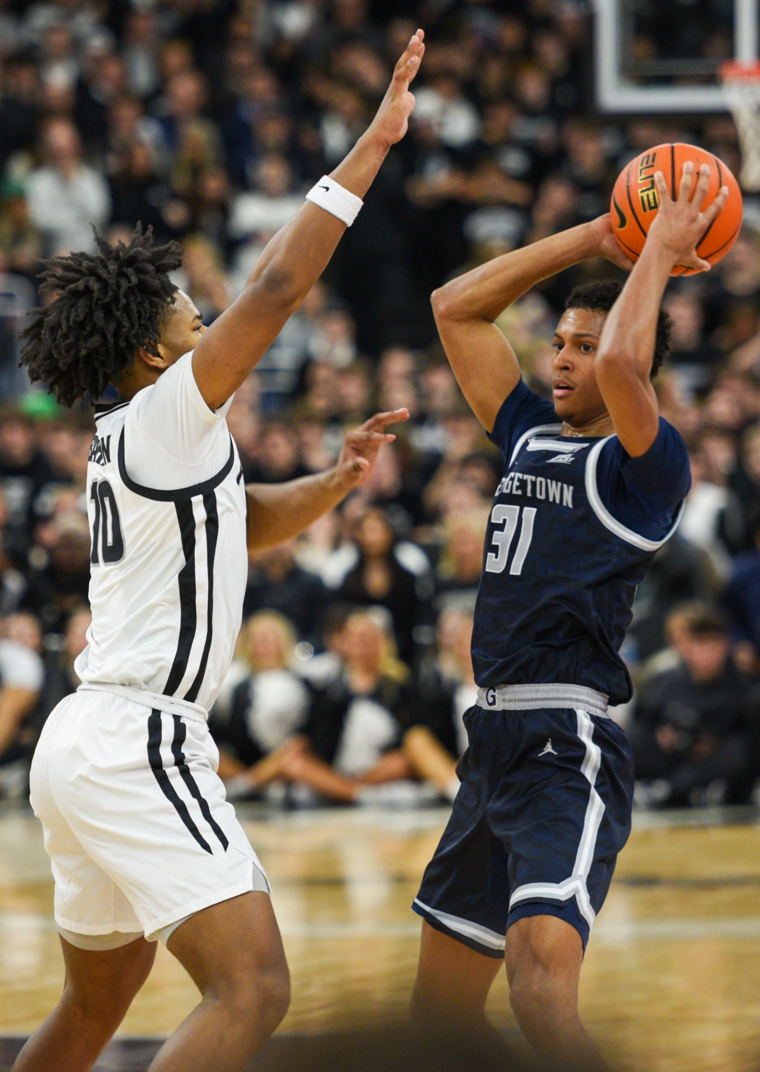 Two basketball players are in the middle of a game. The player on the right wearing a navy blue Georgetown jersey with the number 31 is holding a basketball above his head, preparing to shoot. The player on the left, wearing a white jersey with black