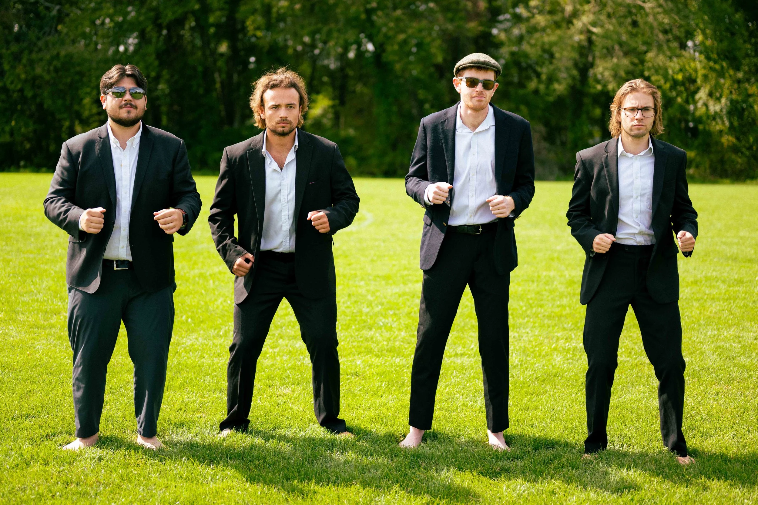 Five men in suits standing barefoot on a green field, with a background of trees, wearing sunglasses, some with their fists clenched.