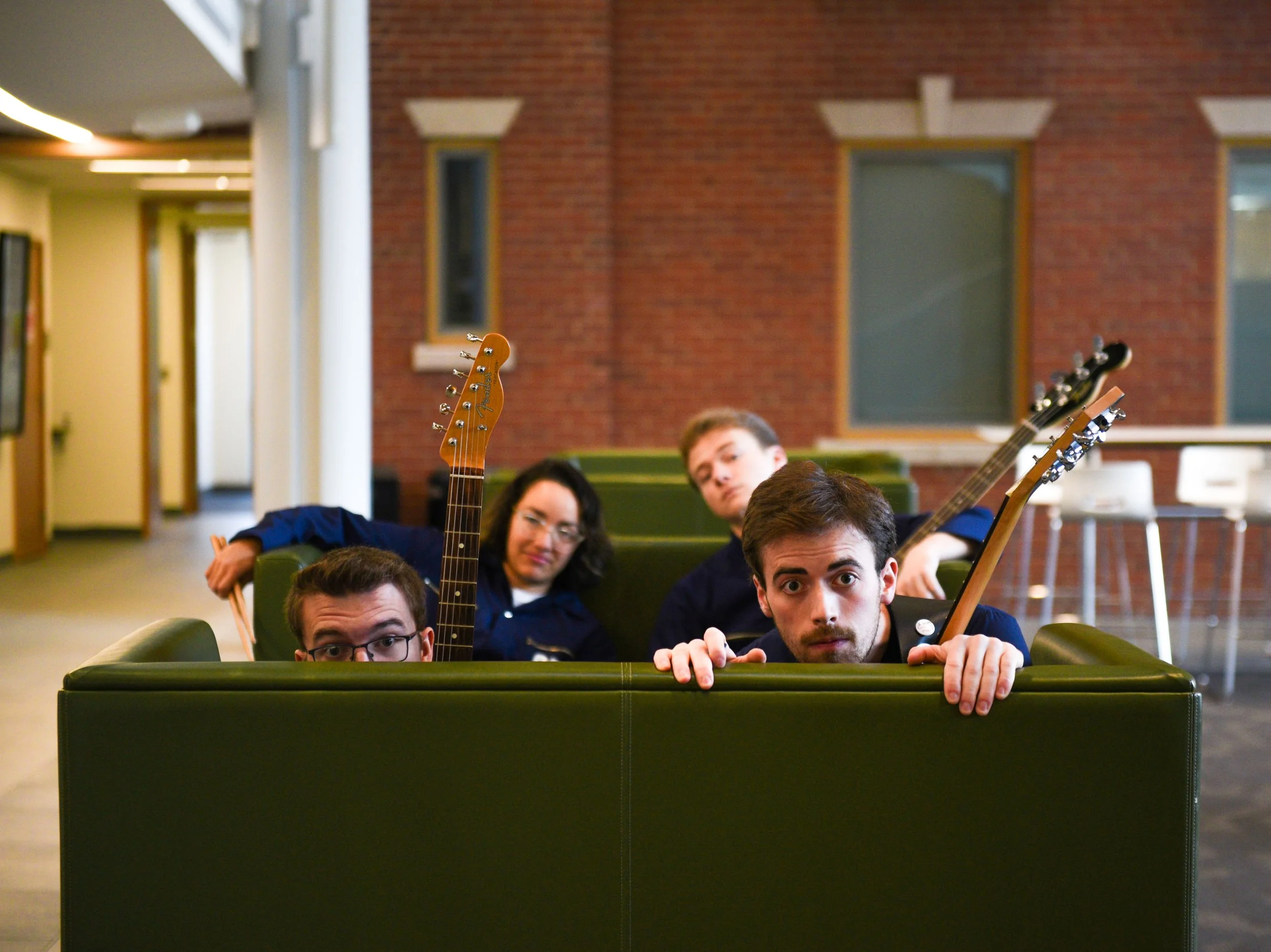 Four people with guitars peeking over the back of a green couch in an indoor lounge area with brick walls and large windows.