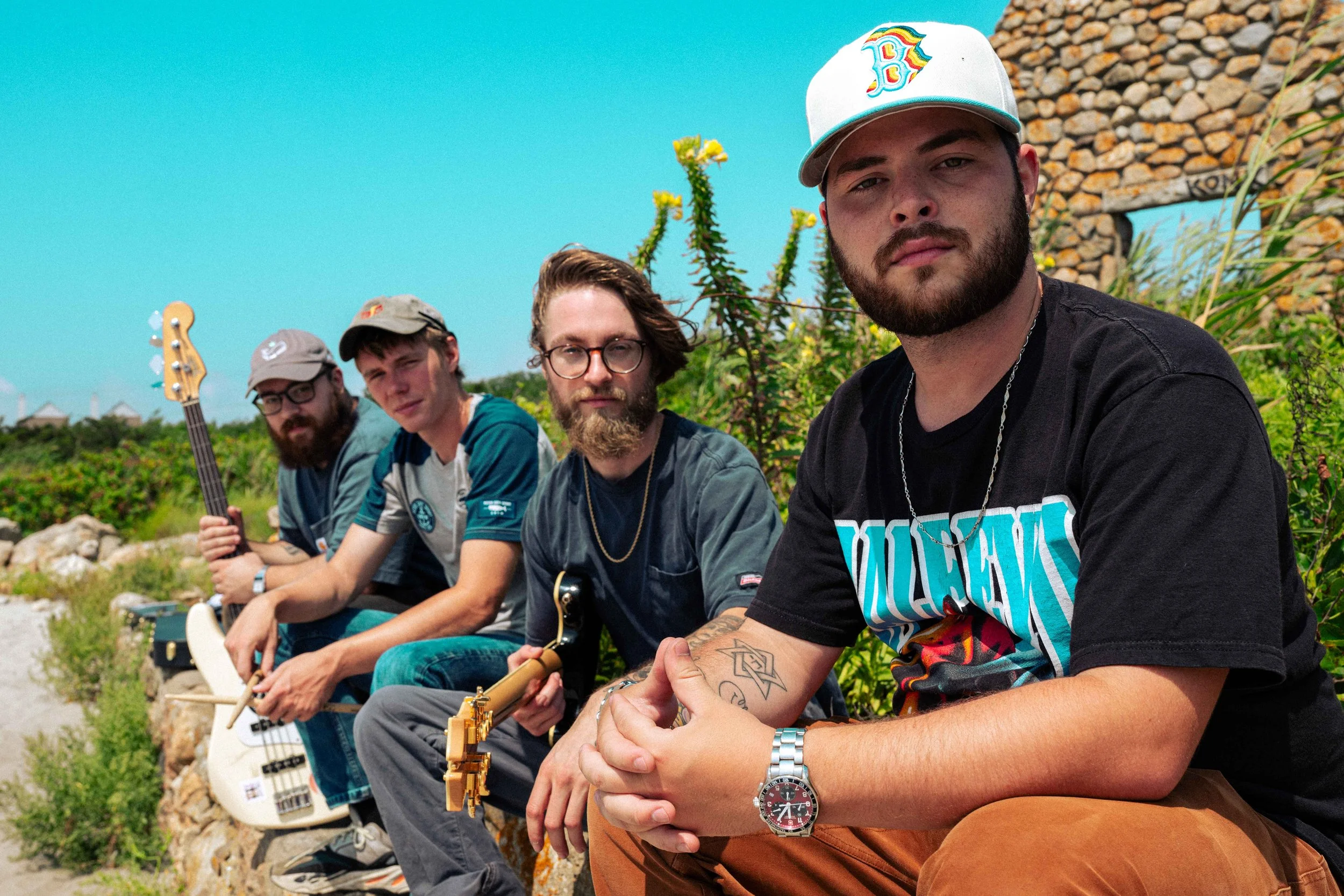 Four men sitting outdoors near a stone wall, holding musical instruments, with green plants and a blue sky in background.
