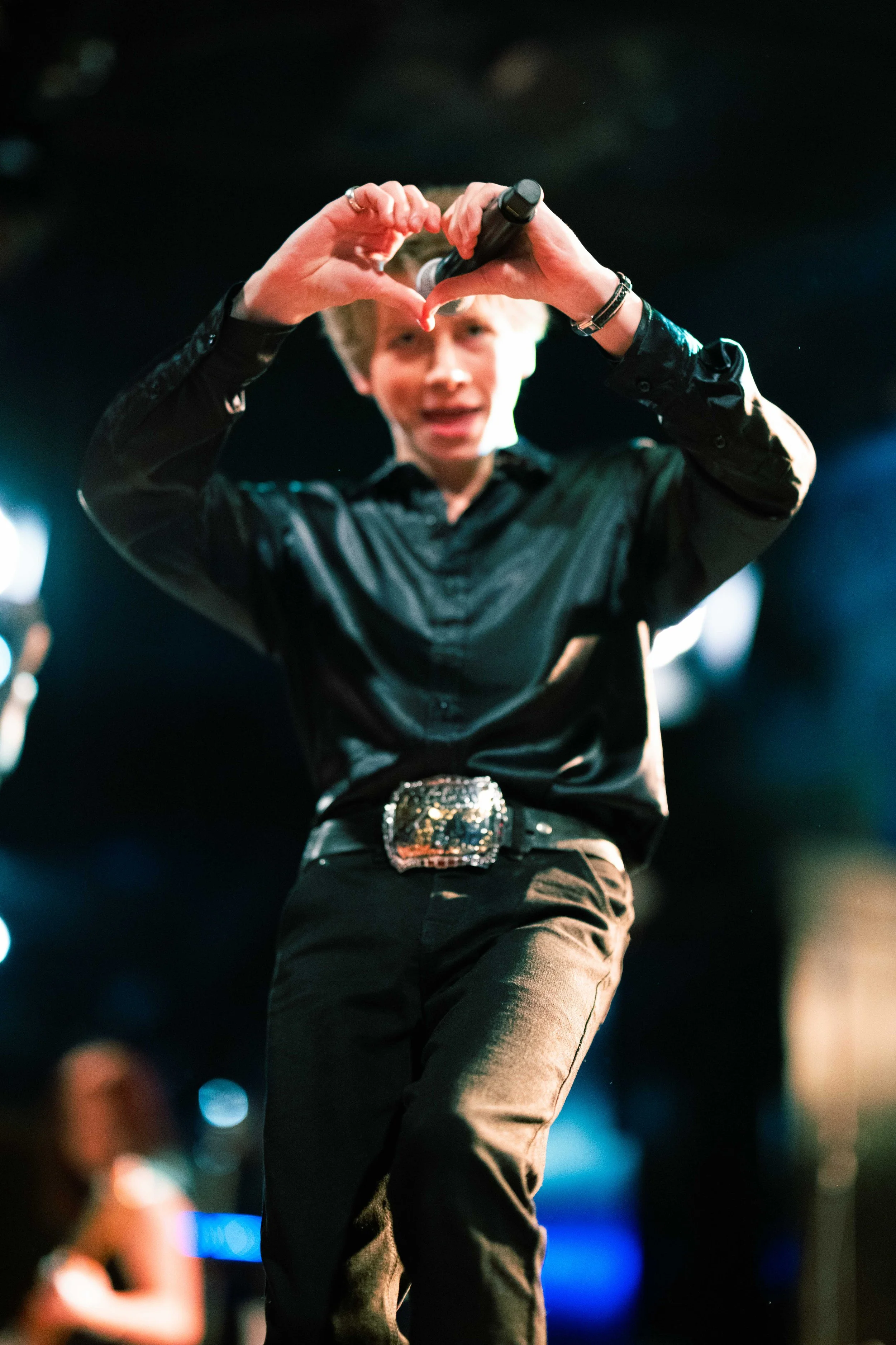 Young person wearing black shirt and pants, making a heart shape with their hands, holding a microphone, in a dimly lit performance setting.