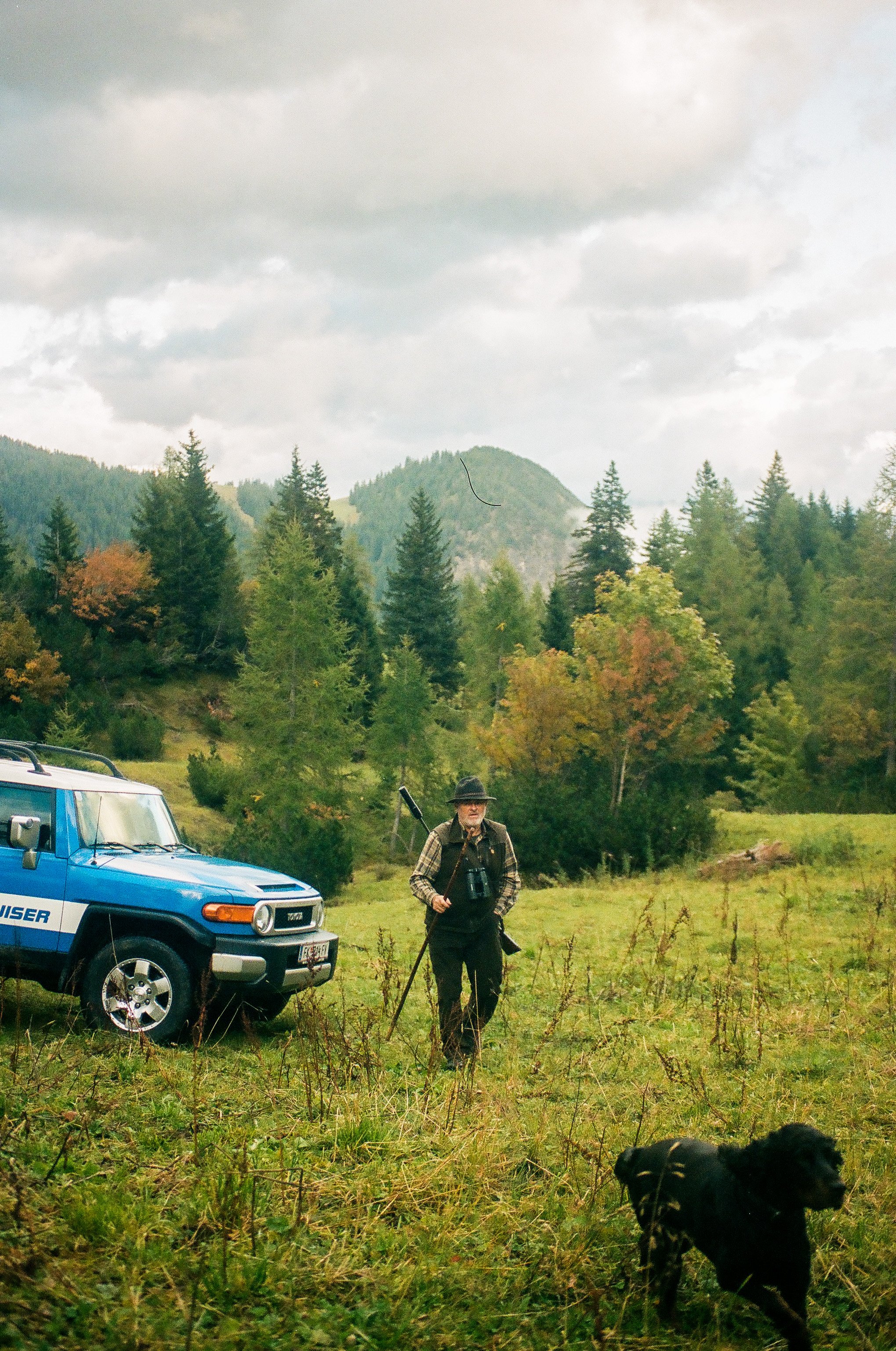 A man stands in a grassy field with a black dog on a leash near a blue off-road vehicle, surrounded by trees and mountains under a cloudy sky.