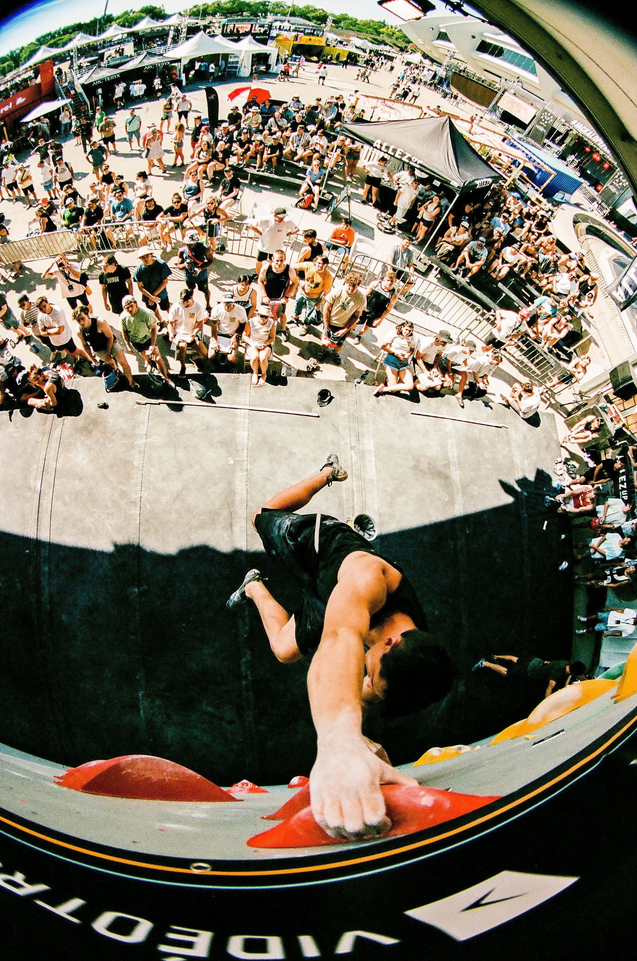 A climber reaching for holds on an indoor rock climbing wall viewed from above, with an outdoor festival or event in the background filled with people, tents, and seating areas.