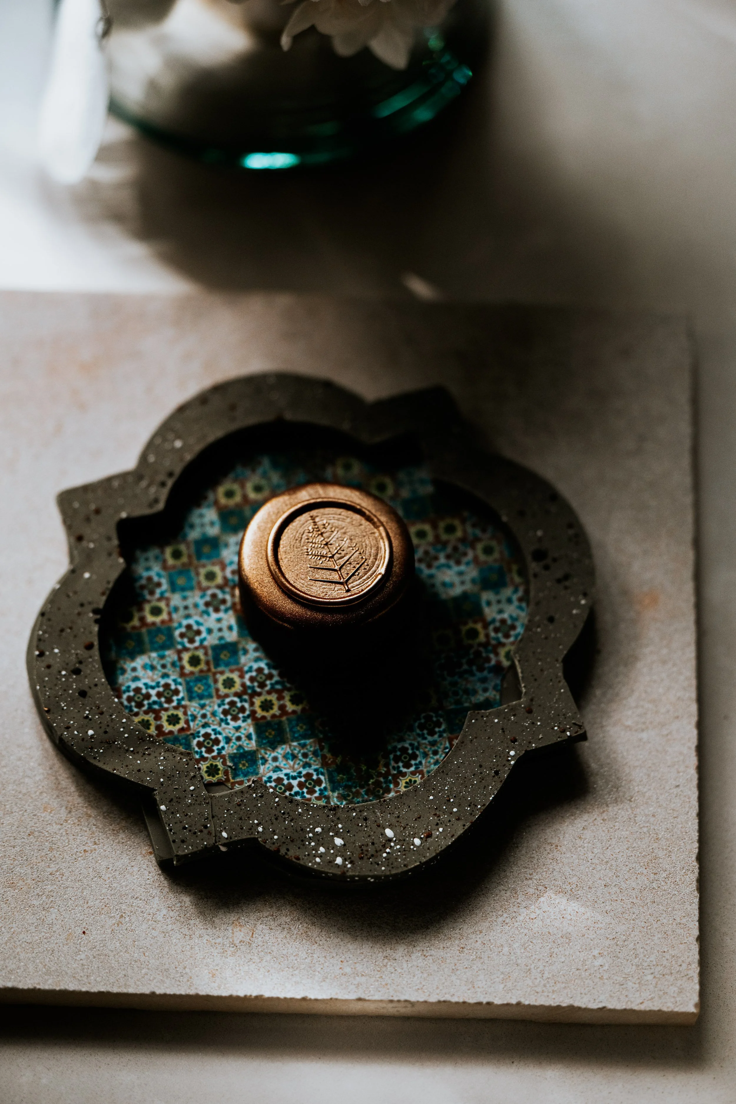 A decorative candle with a leaf design on its top, placed on a patterned ceramic coaster, on a neutral tabletop.