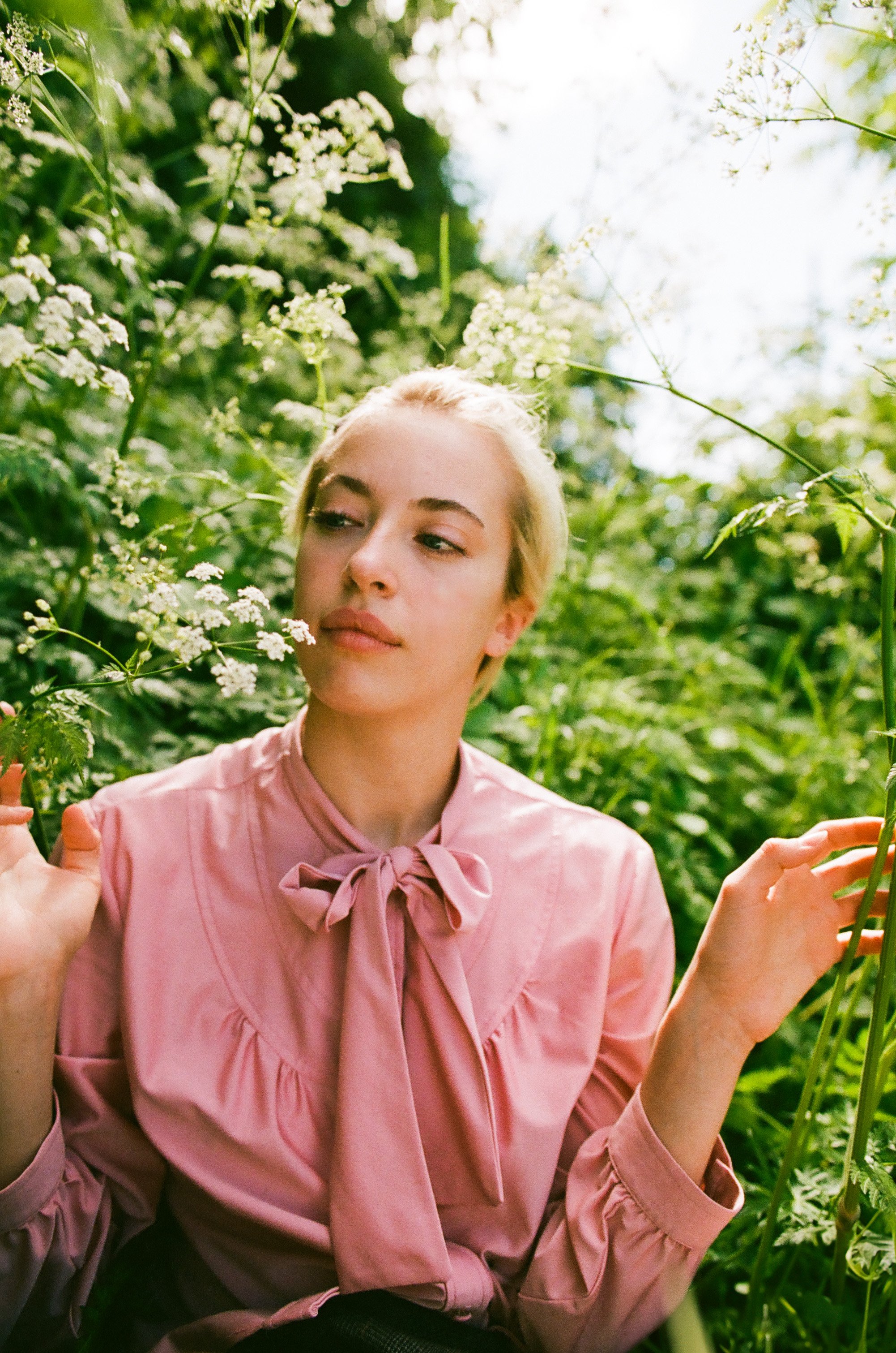 A young woman with blonde hair in a pink blouse with a bow tie, sitting among green foliage and white flowers, looking at the flowers with a serene expression.