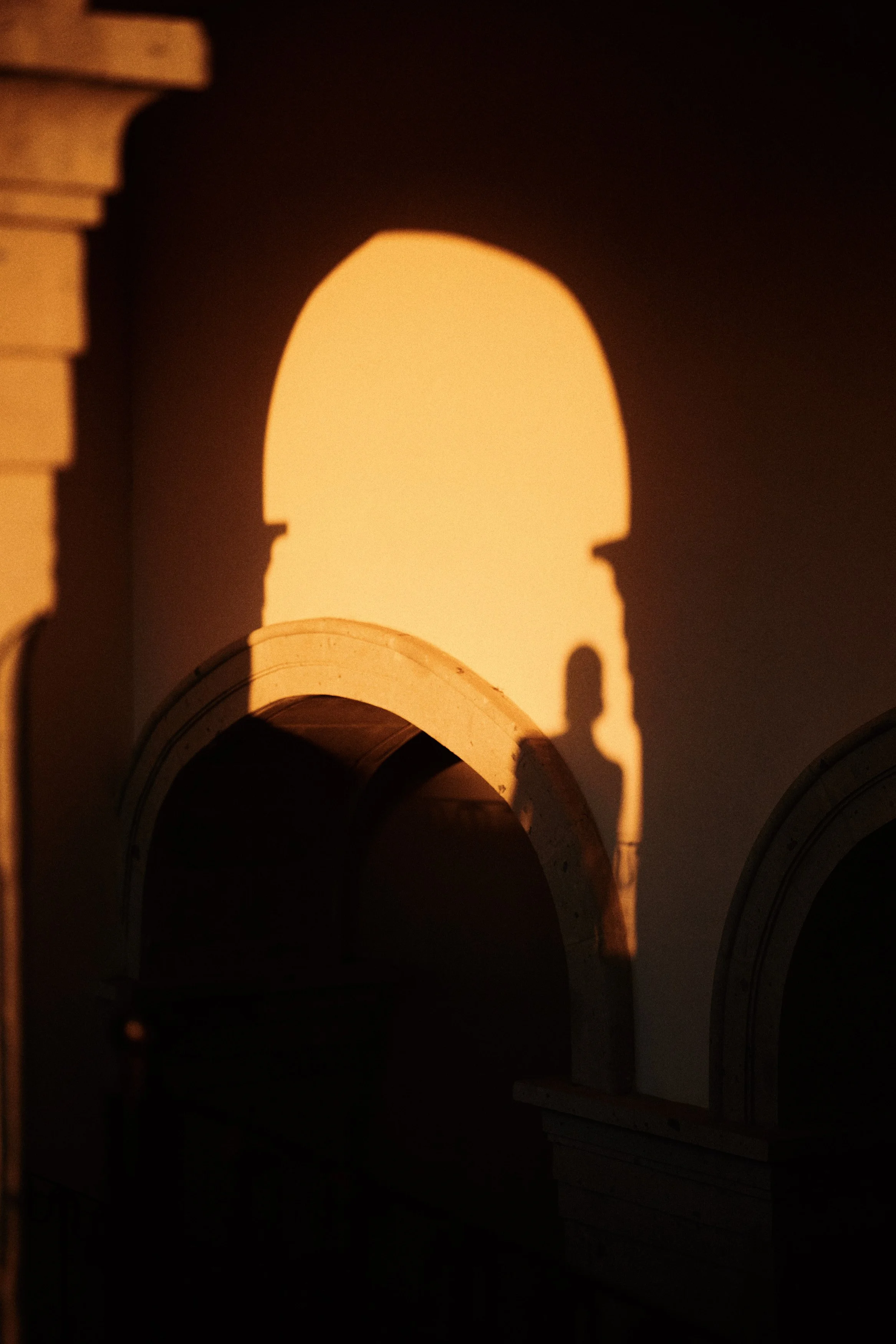 The shadow of a person cast on a wall created by the setting sun, with an archway visible in the foreground and warm lighting.