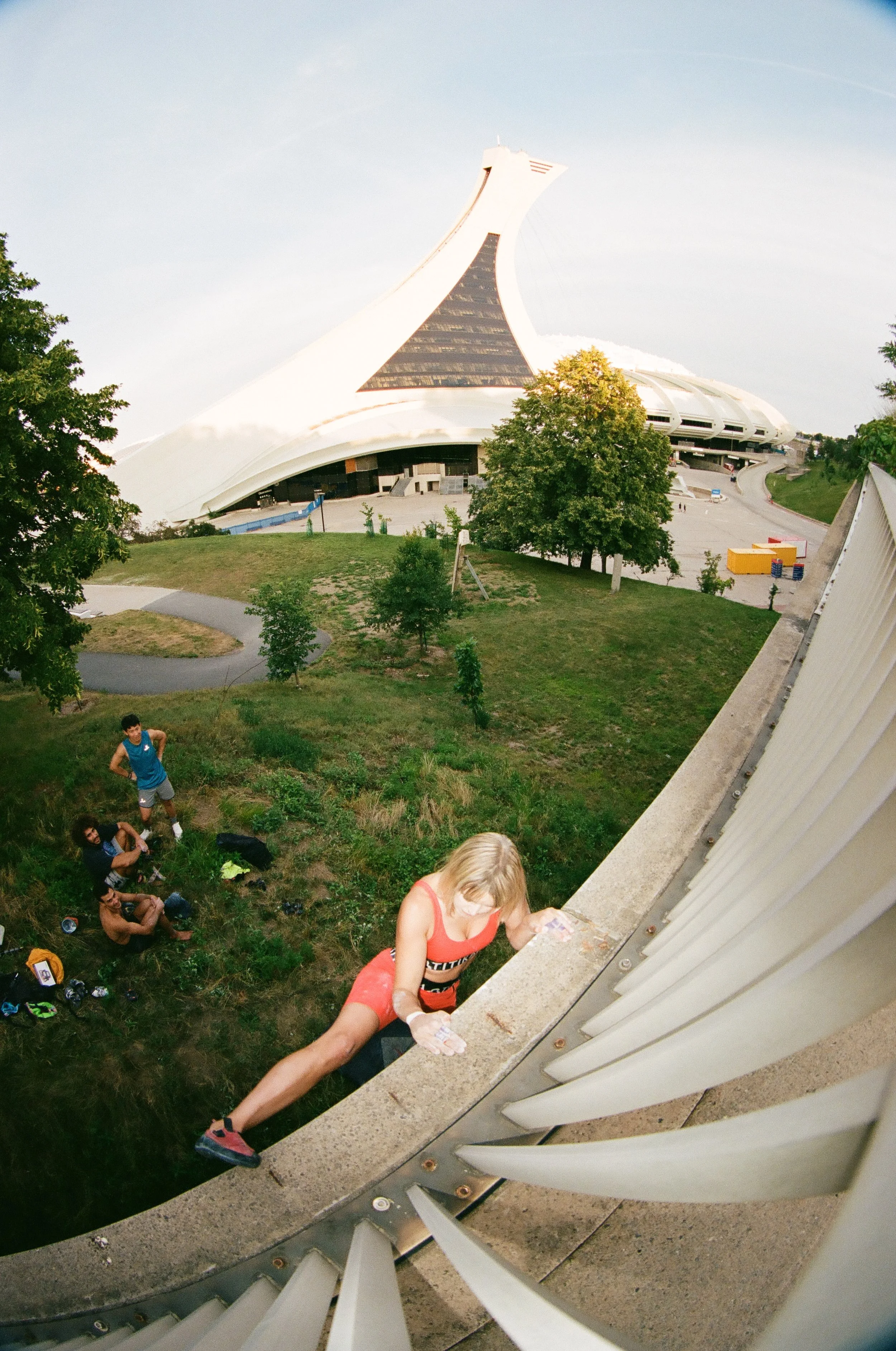 A woman climbing a concrete ledge with a modern, alien-like building in the background, and two people sitting on the grass below.