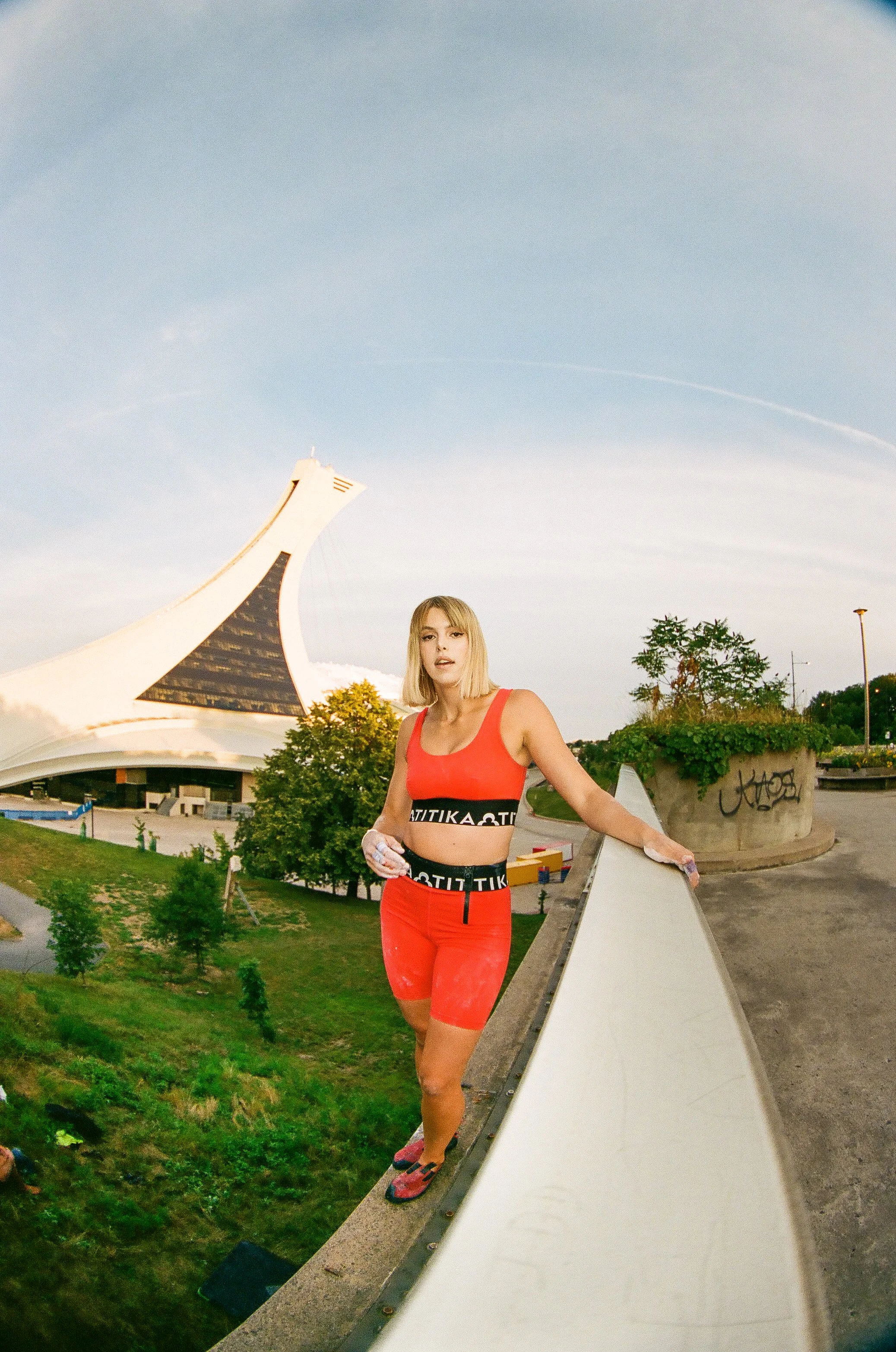 A woman in red athletic wear standing on a rooftop ledge with a modern architectural building in the background.