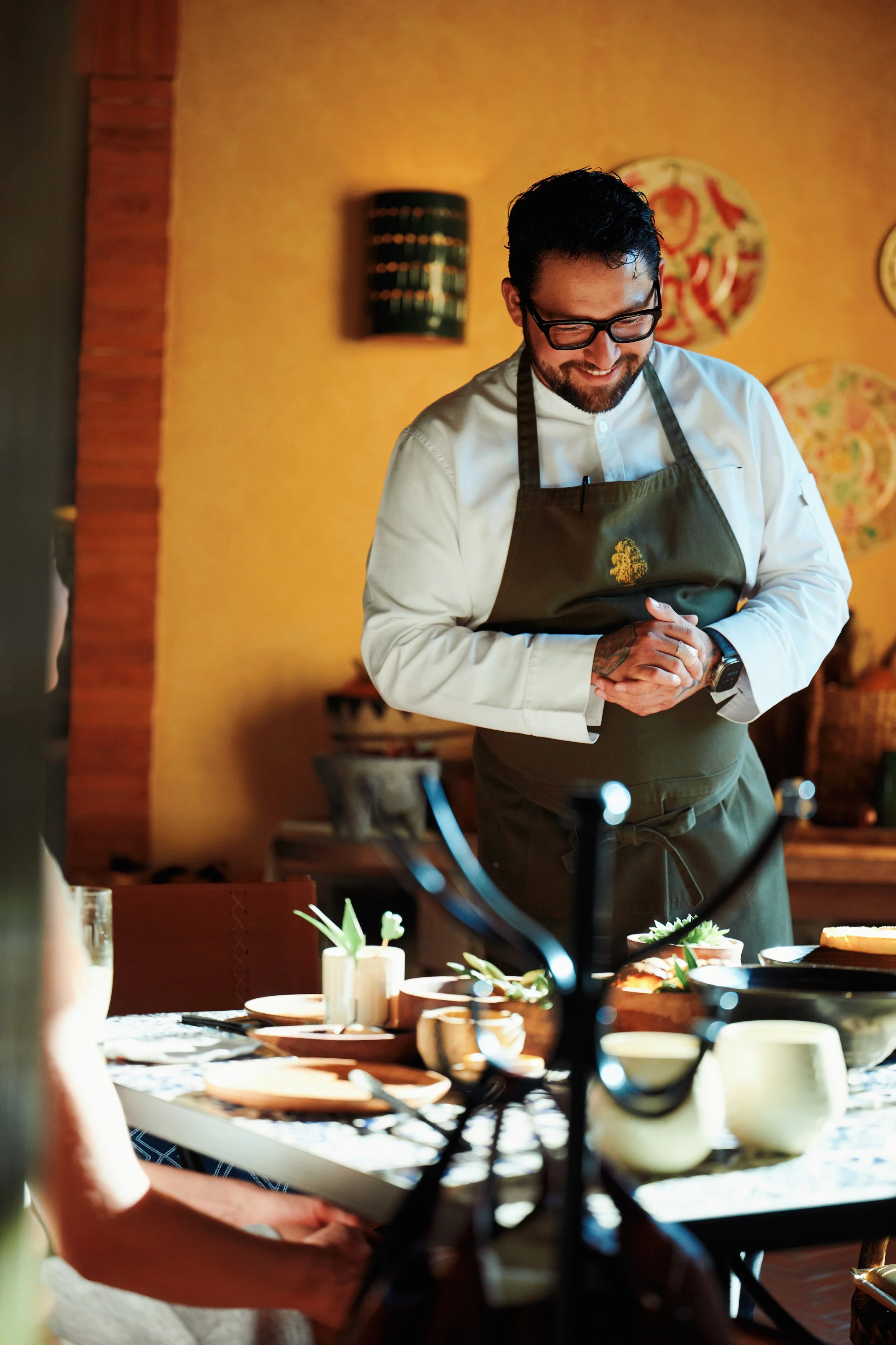 A smiling male chef wearing glasses and a white apron standing in a warmly lit restaurant kitchen or dining area, with a table set with dishes and utensils in front of him.