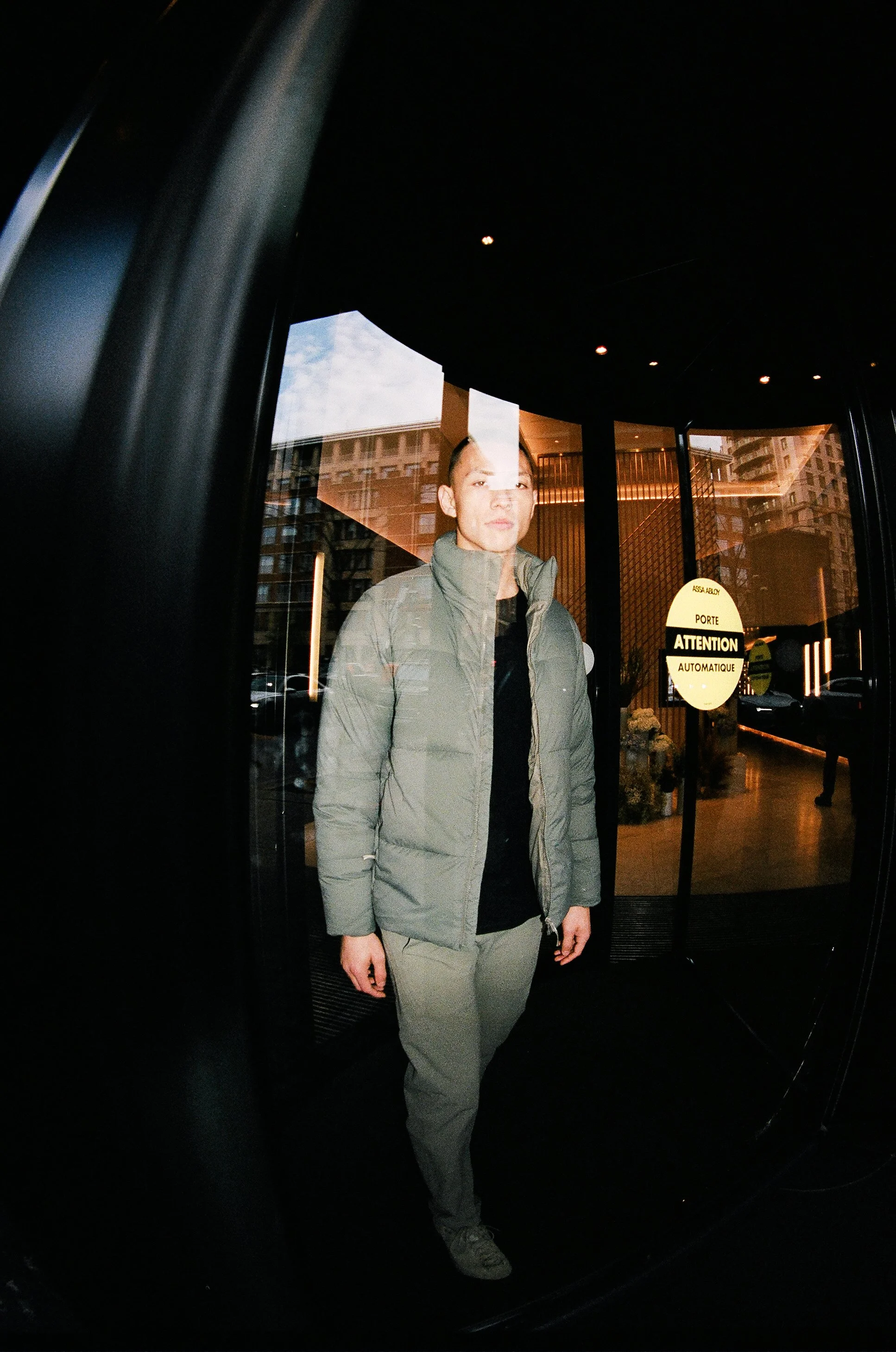 A man wearing a gray jacket and pants standing behind a glass door with signs that read 'Attention Automatique' and 'Porte.' Reflection of buildings and the sky can be seen in the glass.