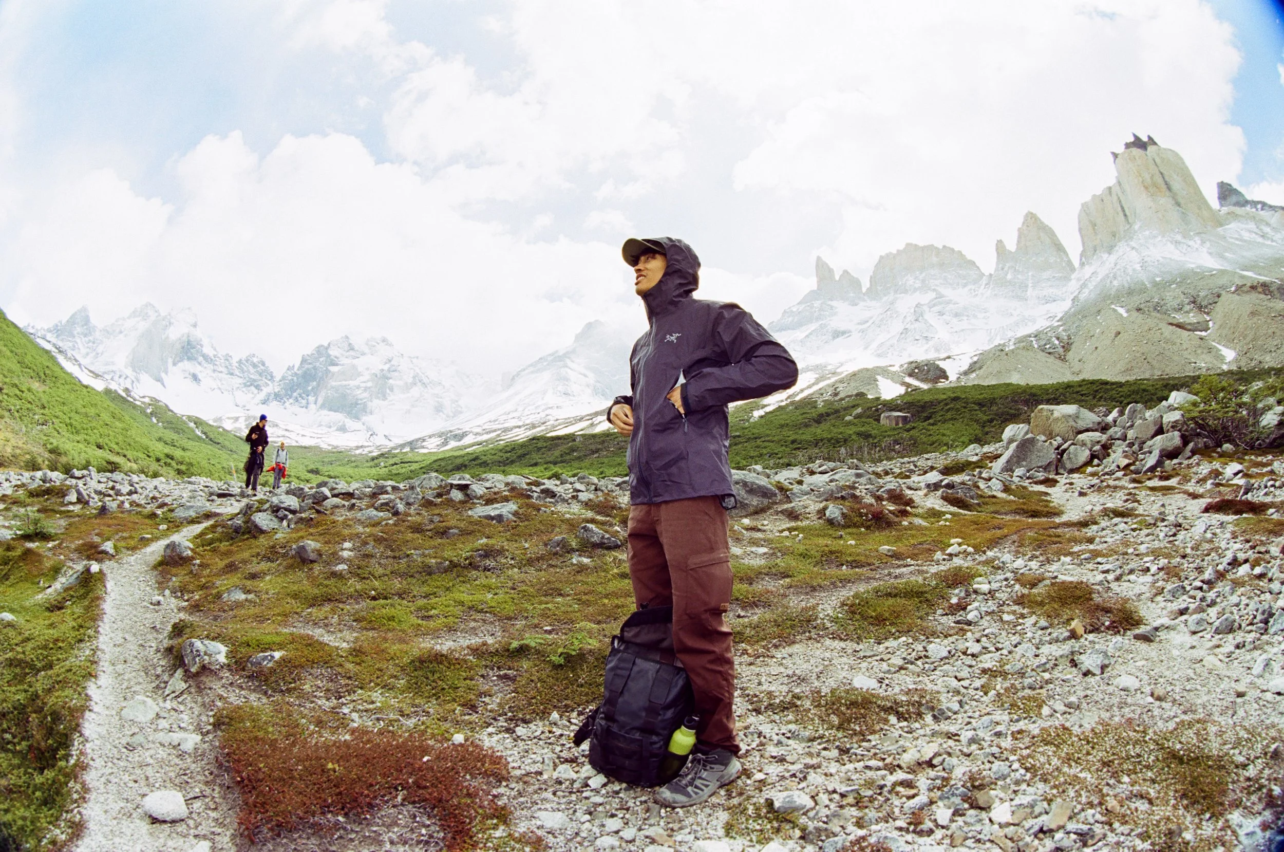 A woman in a black rain jacket and brown pants standing on a rocky trail in a mountainous area with snow-capped peaks in the background, and two people visible further down the trail.