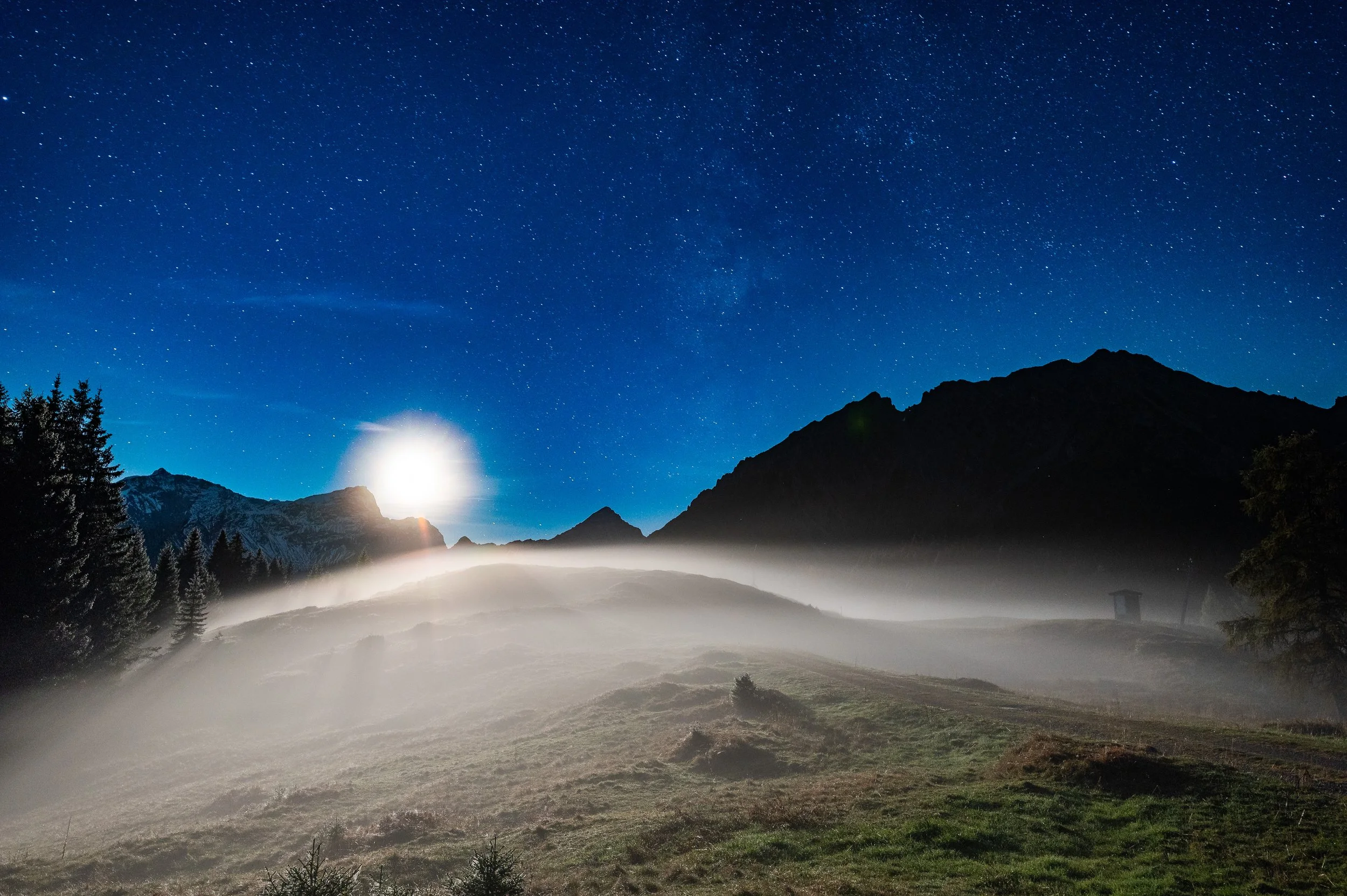 Nighttime mountain landscape with a bright moon, stars, fog over grassy hills, and silhouette of trees and mountains.