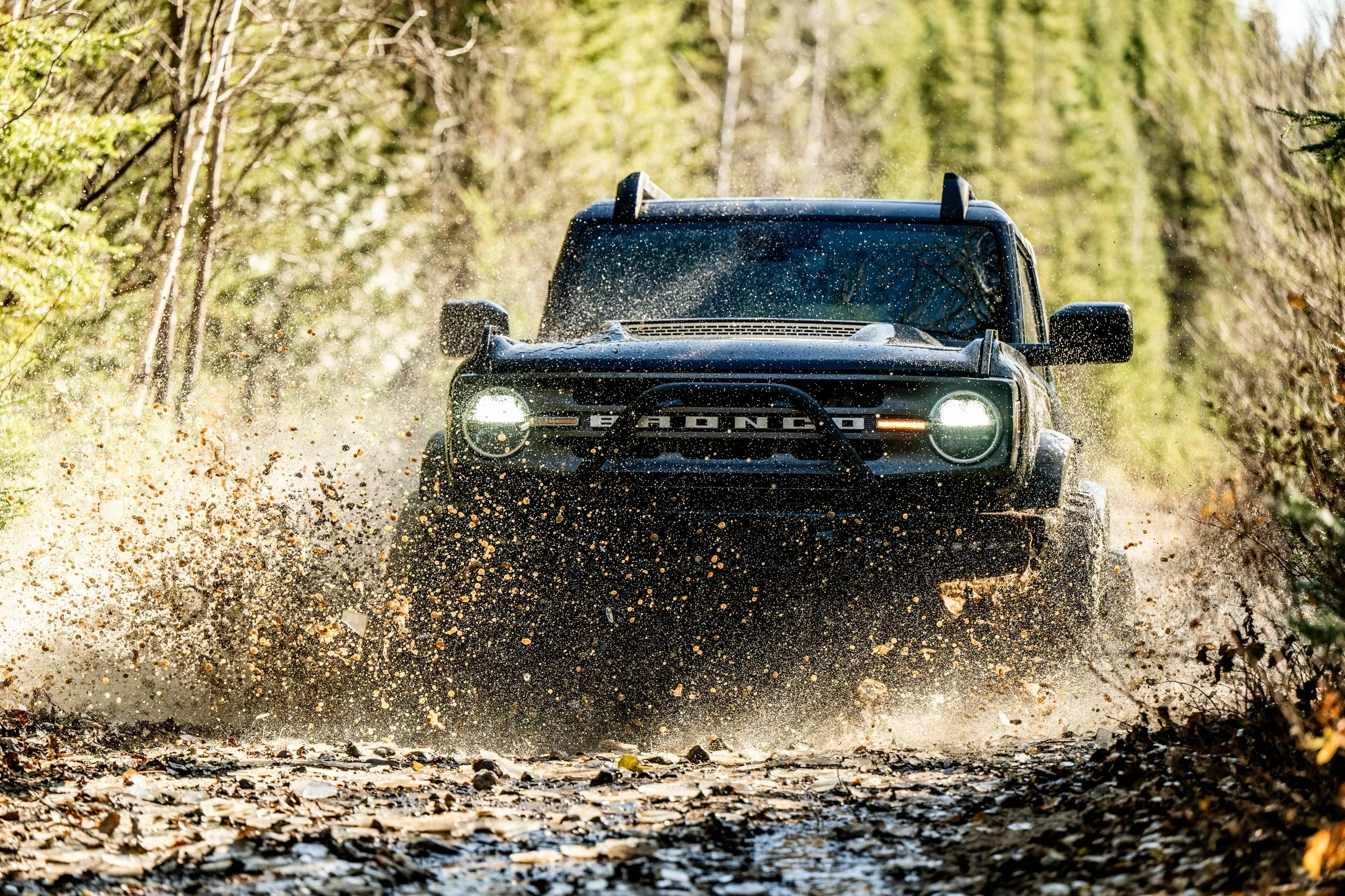 A black Land Rover Defender driving through a muddy forest trail with splashing mud and leaves, surrounded by green trees.