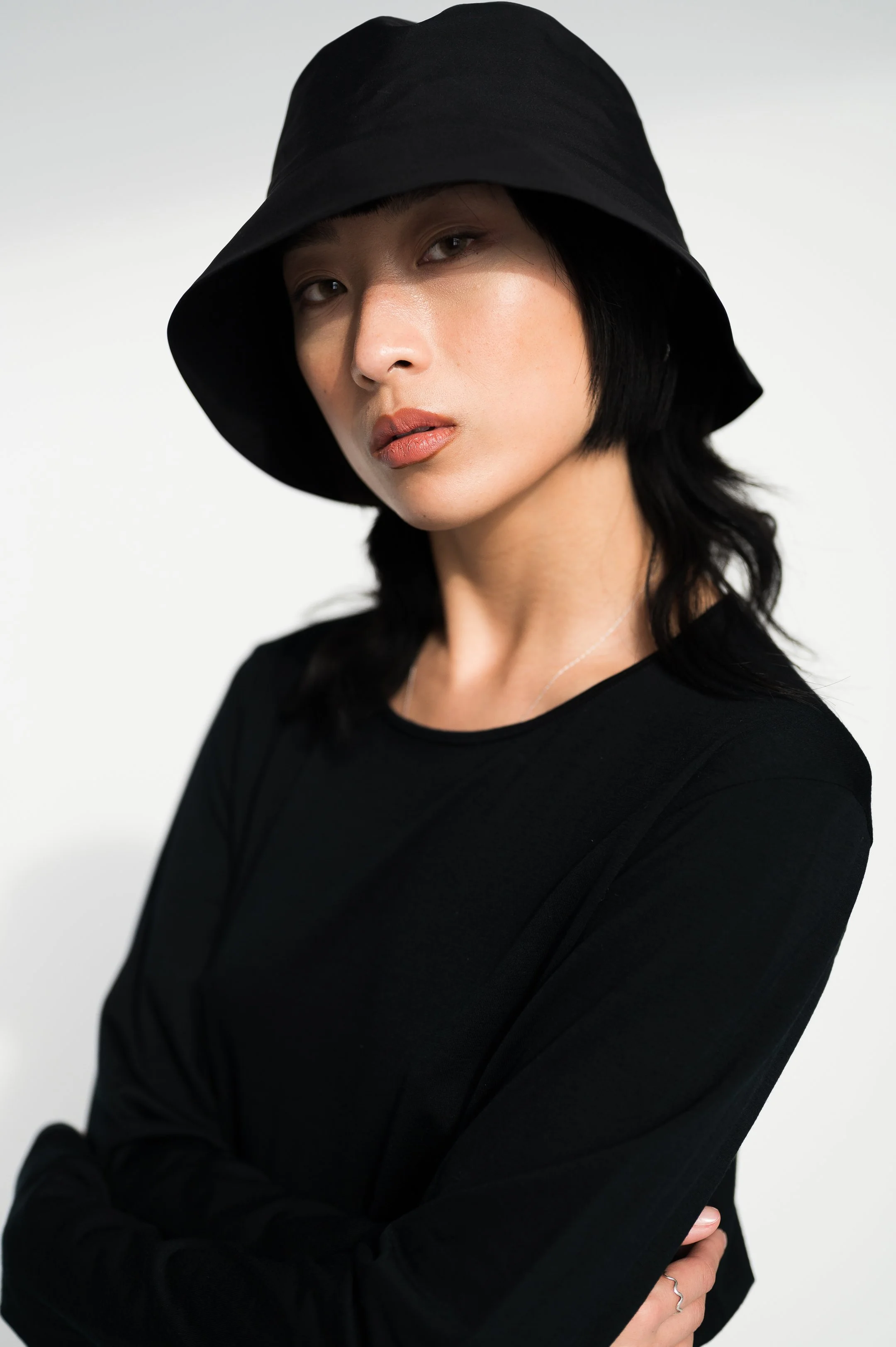 A woman with straight black hair, wearing a black wide-brimmed hat and black long-sleeve shirt, posing with her arms crossed against a plain white background.