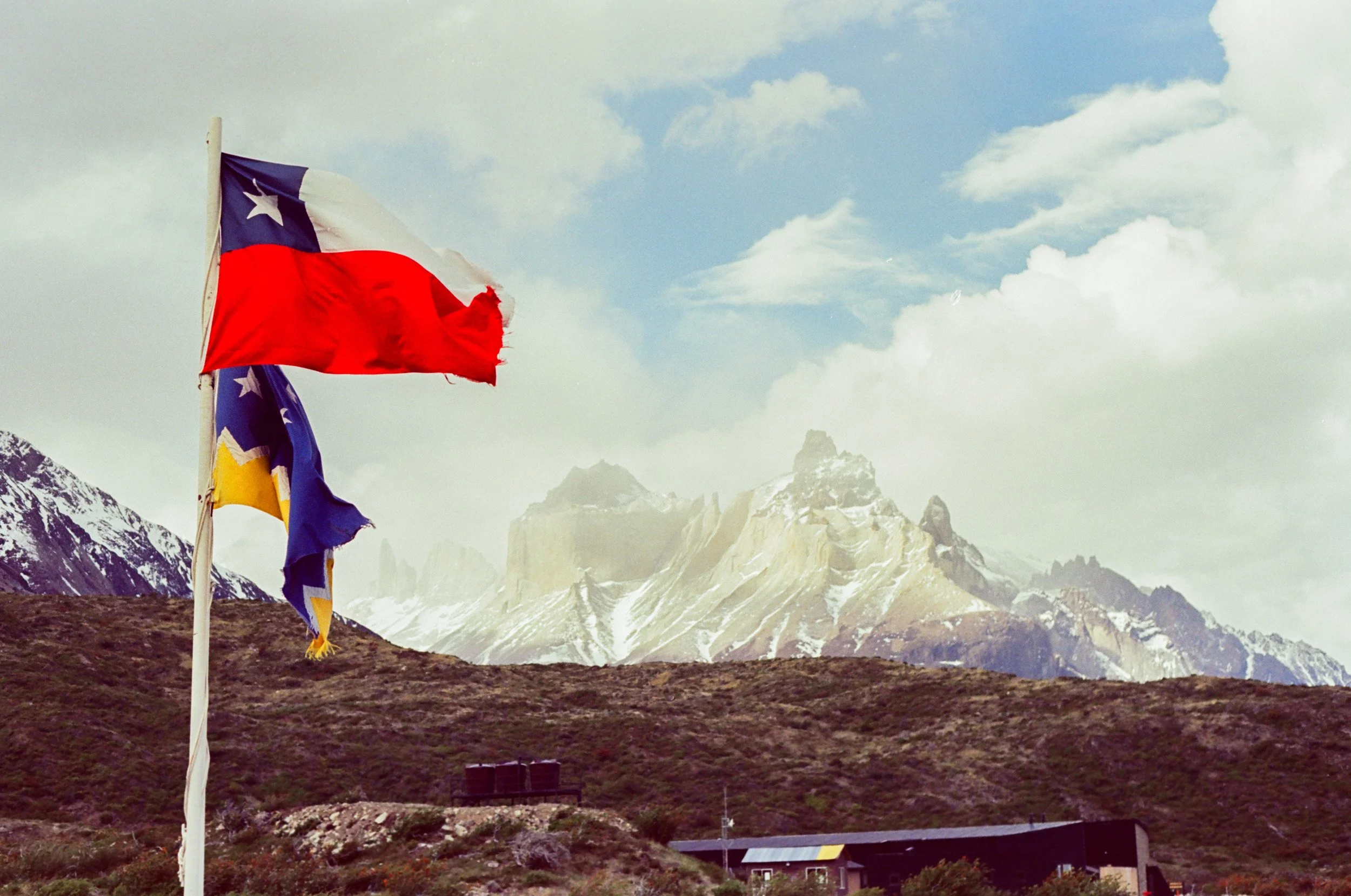 Chilean flags waving in front of the Andes Mountains with snow-capped peaks and a partly cloudy sky.