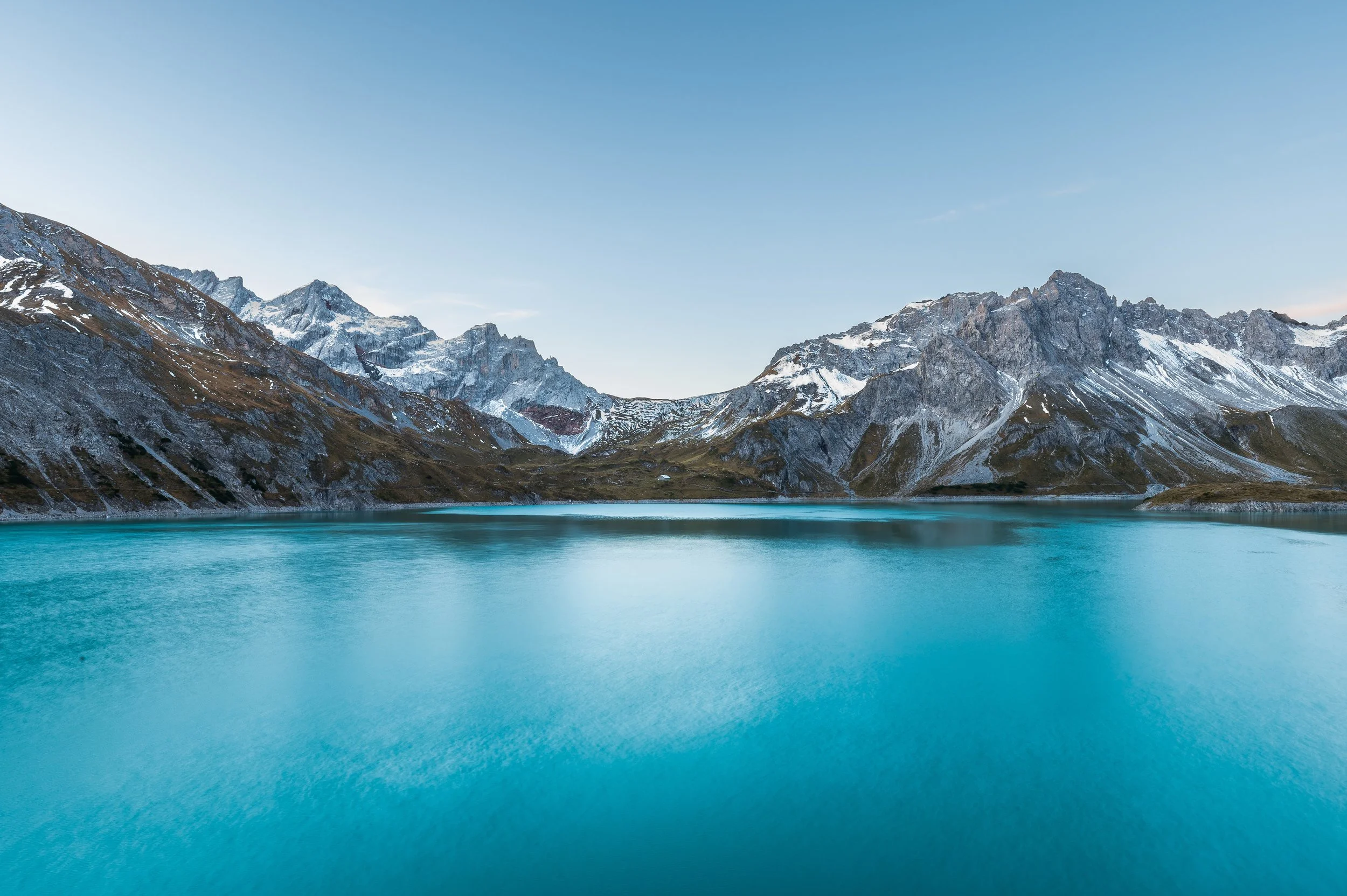 A serene mountain lake with turquoise water, surrounded by snow-capped rugged mountains under a clear blue sky.