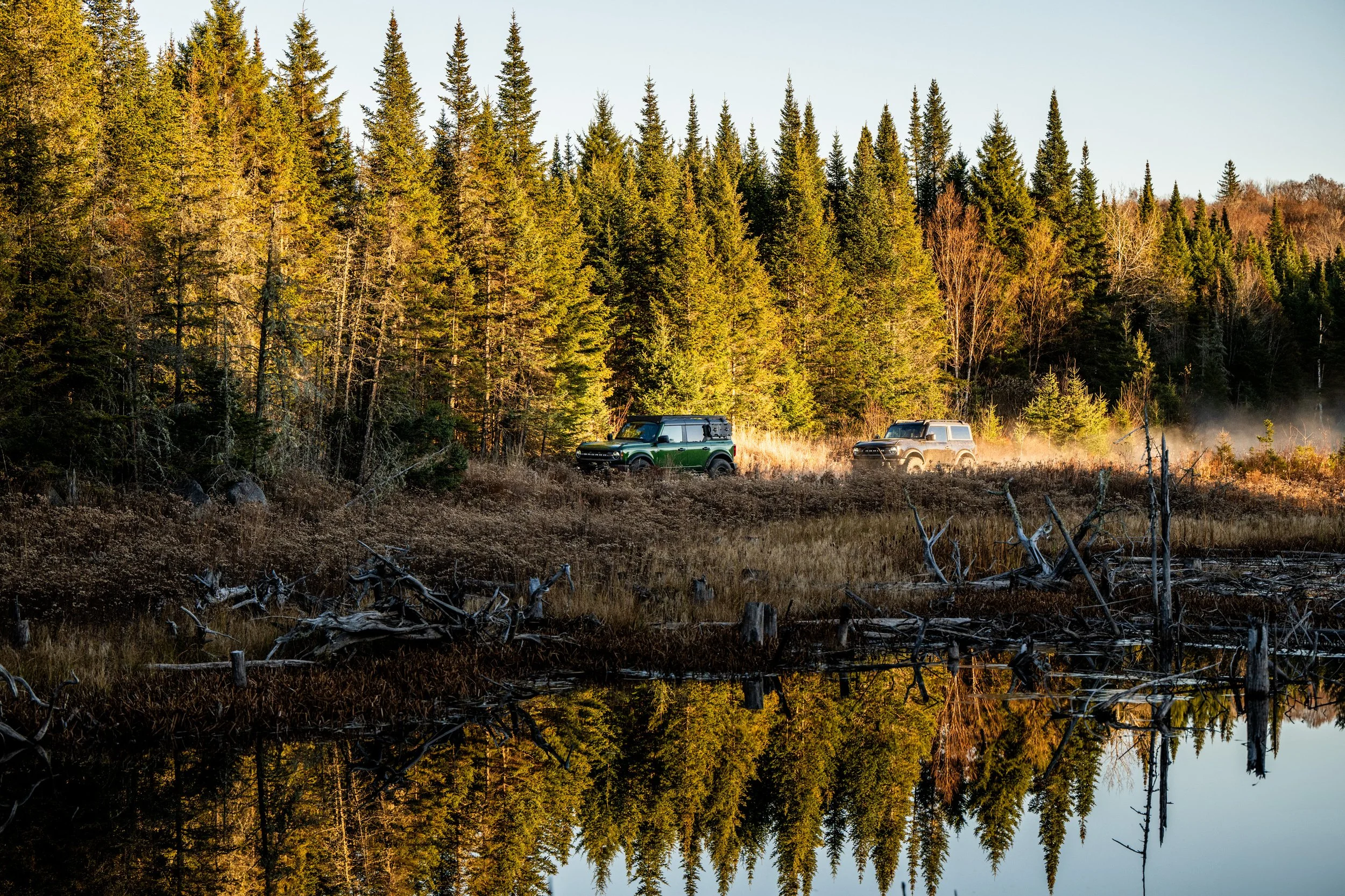 Two off-road vehicles driving through a forest area near a body of water, with trees and mist in the background.
