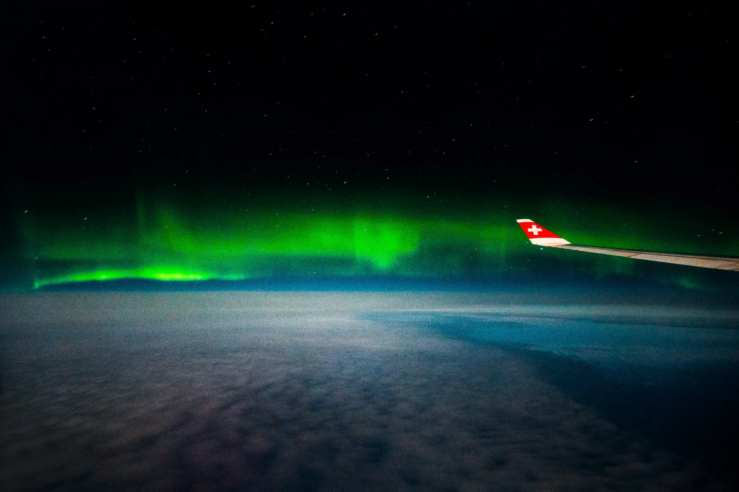 Nighttime view from an airplane window showing the northern lights (aurora borealis) above clouds with a plane's wingtip on the right side, marked with a Swiss Swissair logo.