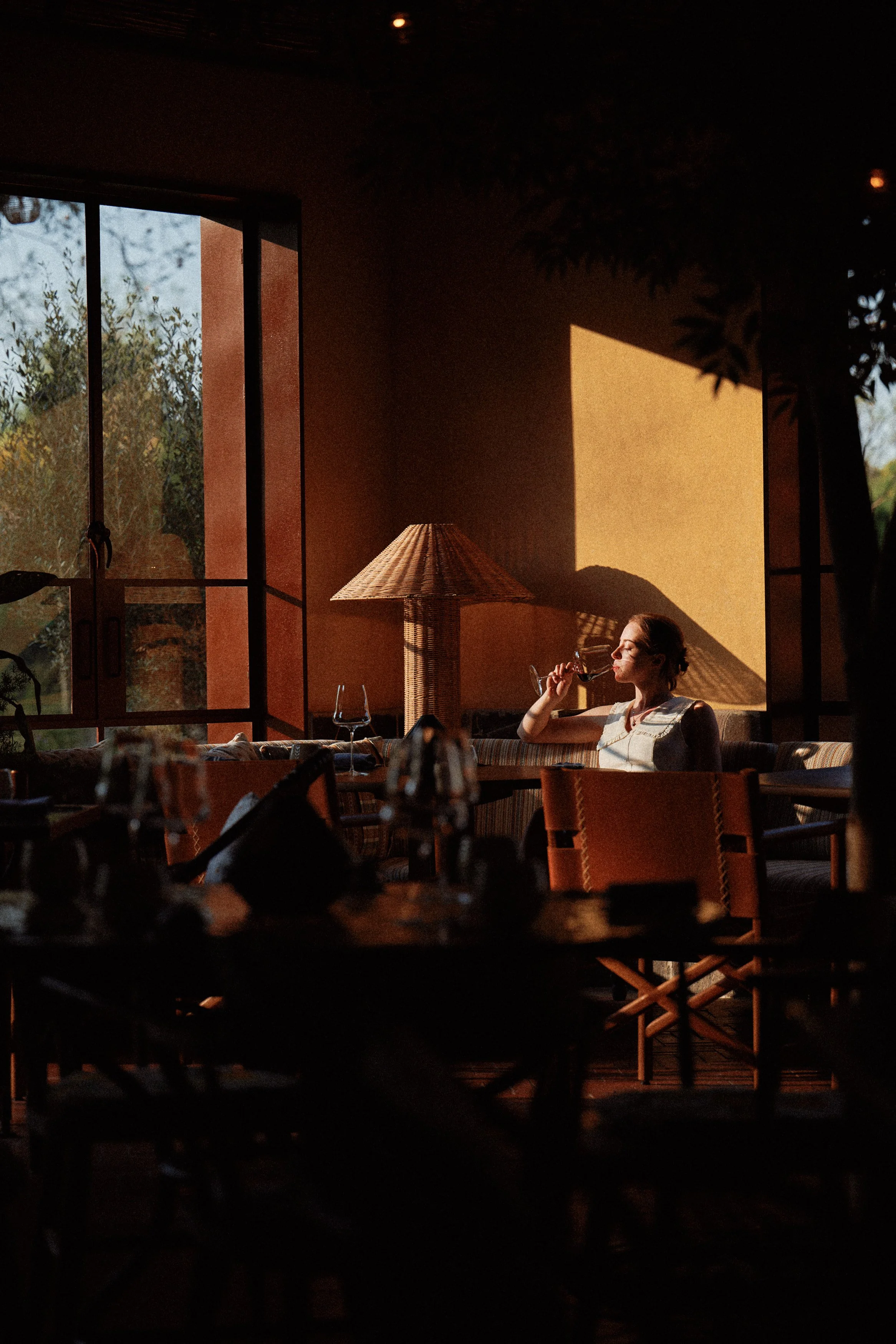 A woman sitting in a warmly lit restaurant, drinking wine, with sunlight casting a shadow on the wall behind her.