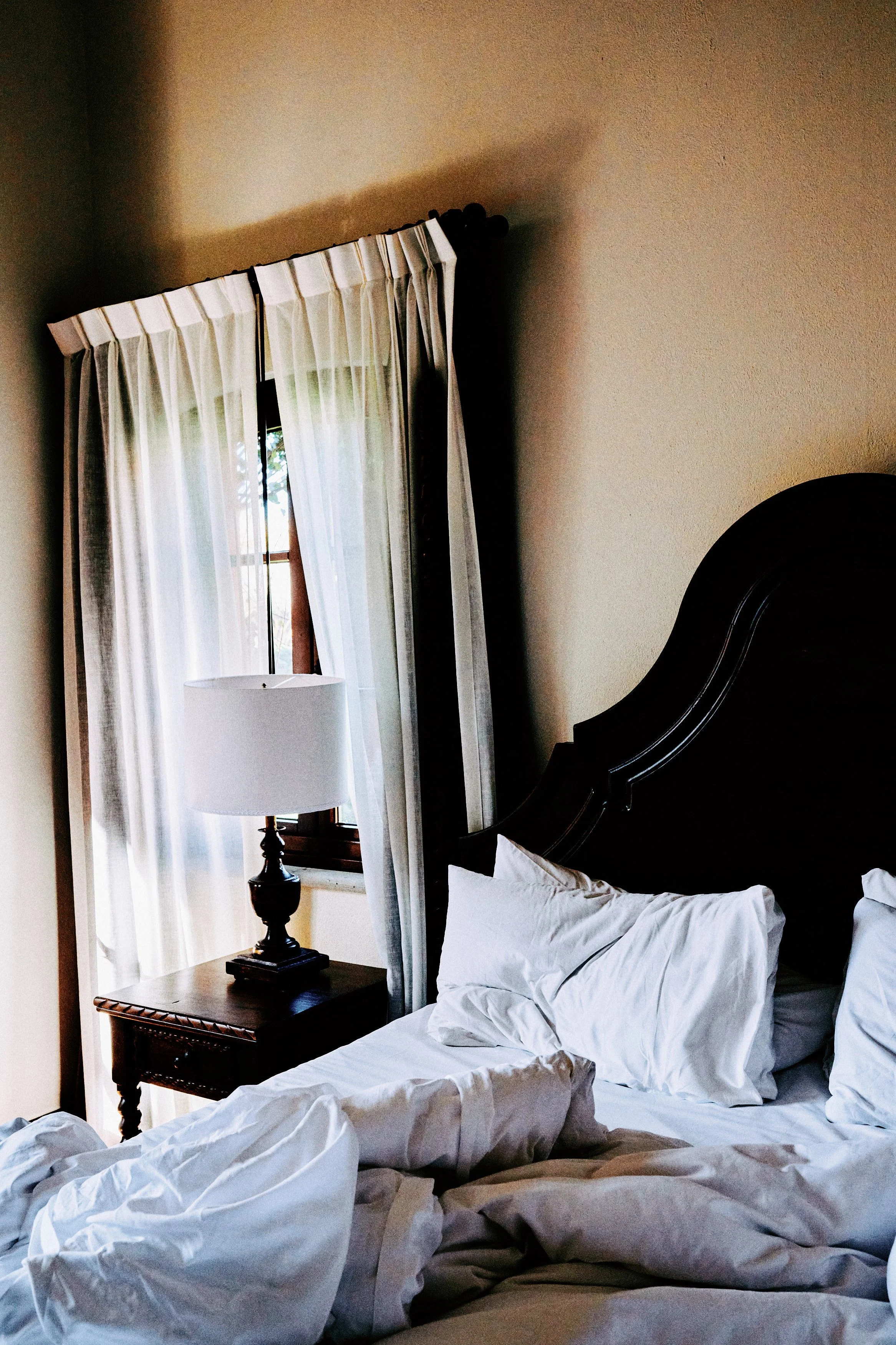 A bedroom scene with a disheveled bed, a wooden nightstand with a lamp, and a window with white curtains that are partly open letting in natural light.