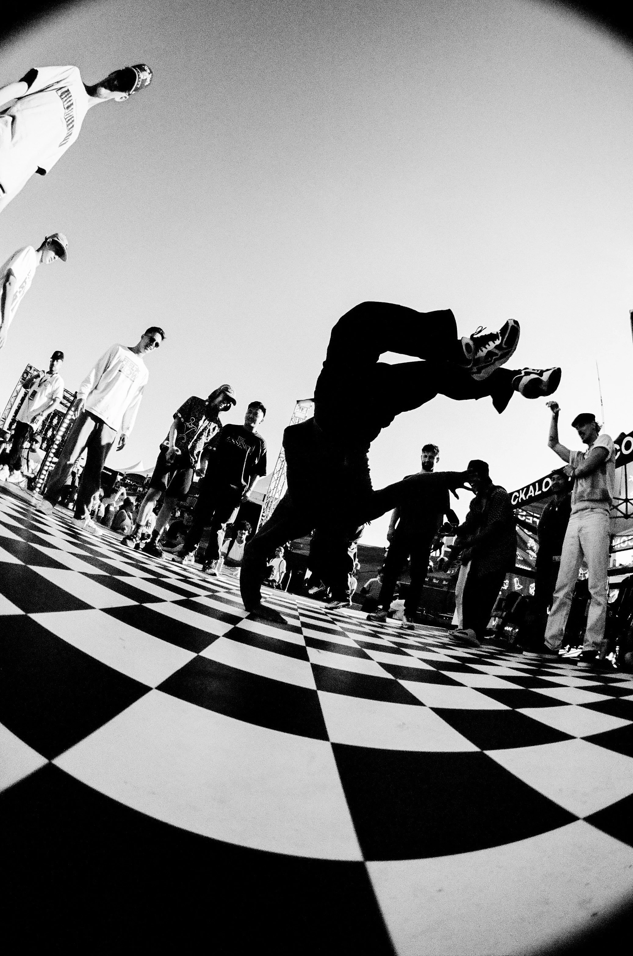 A person breaks dancing on a checkered floor with a crowd watching around, shot in black and white.