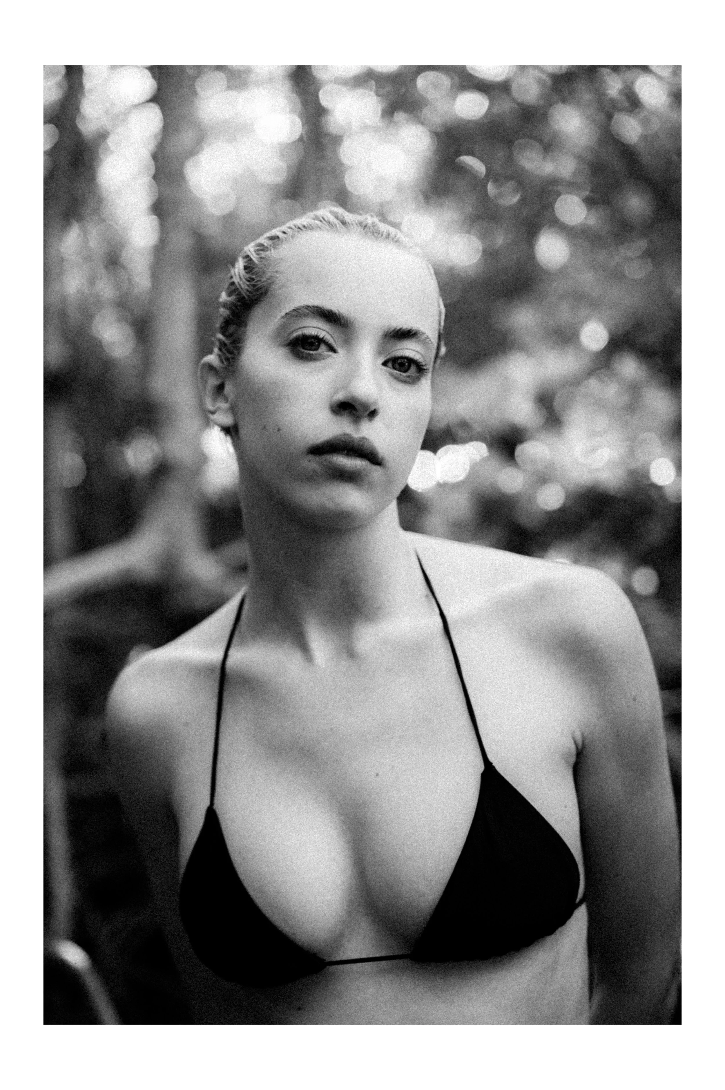 Black and white photograph of a woman with braided hair, wearing a bikini top, standing outdoors with blurred trees in the background.