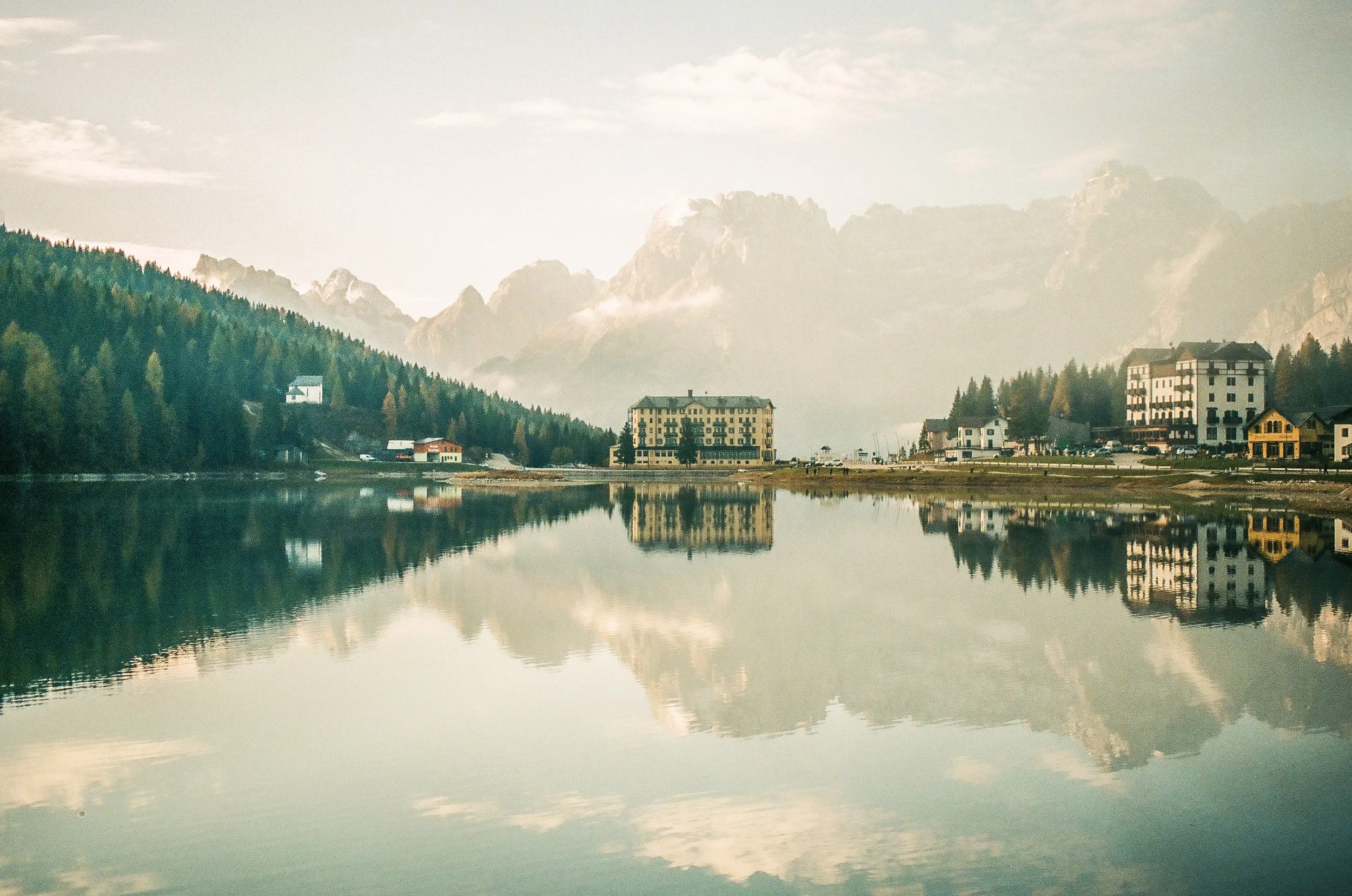 A calm lake with reflections of buildings, trees, and mountains in the water, under a cloudy sky.