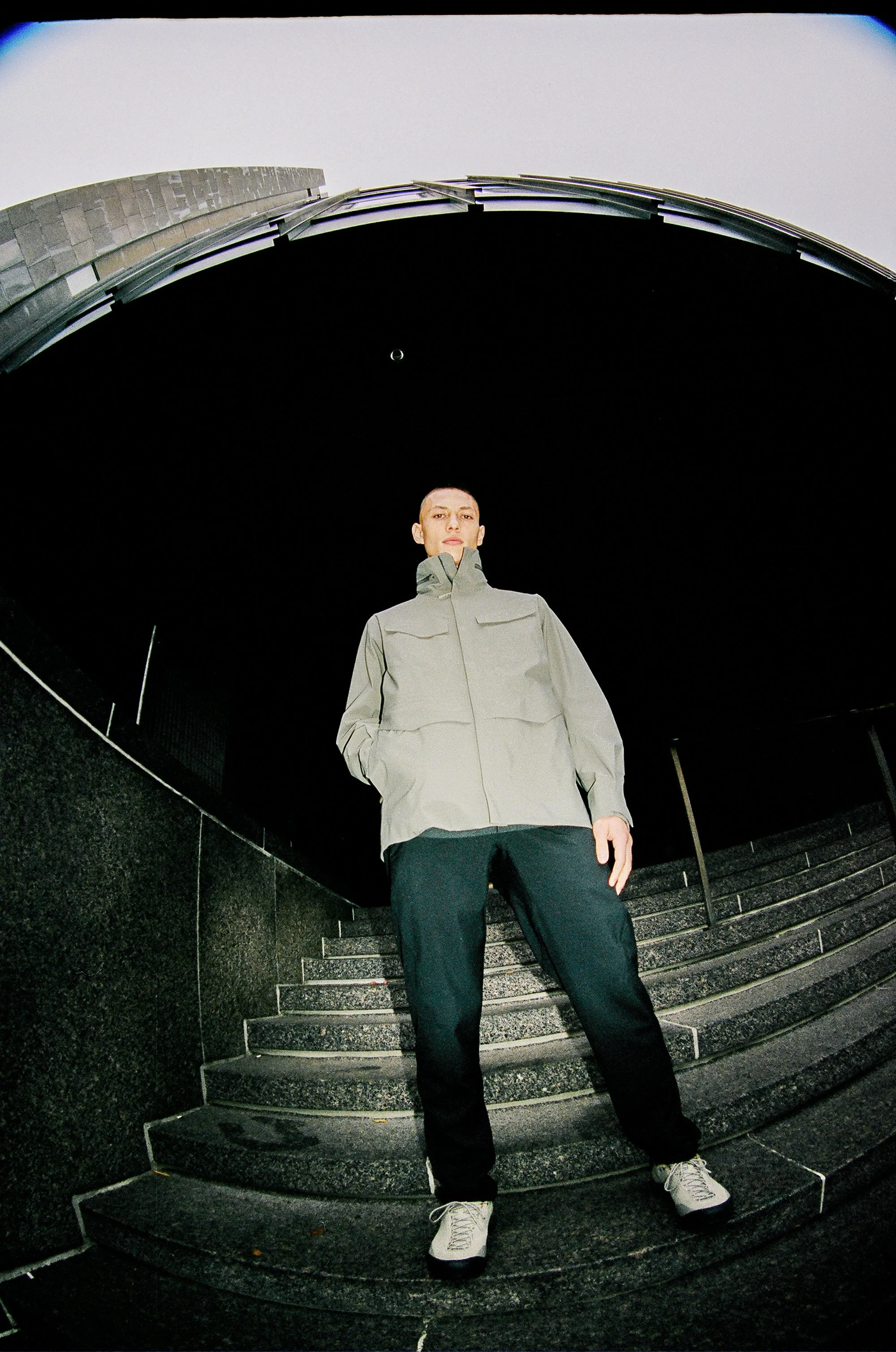 Person standing on stairs outdoors at night, wearing a gray jacket, black pants, and white shoes, with a dark cityscape background and a distorted fisheye effect.