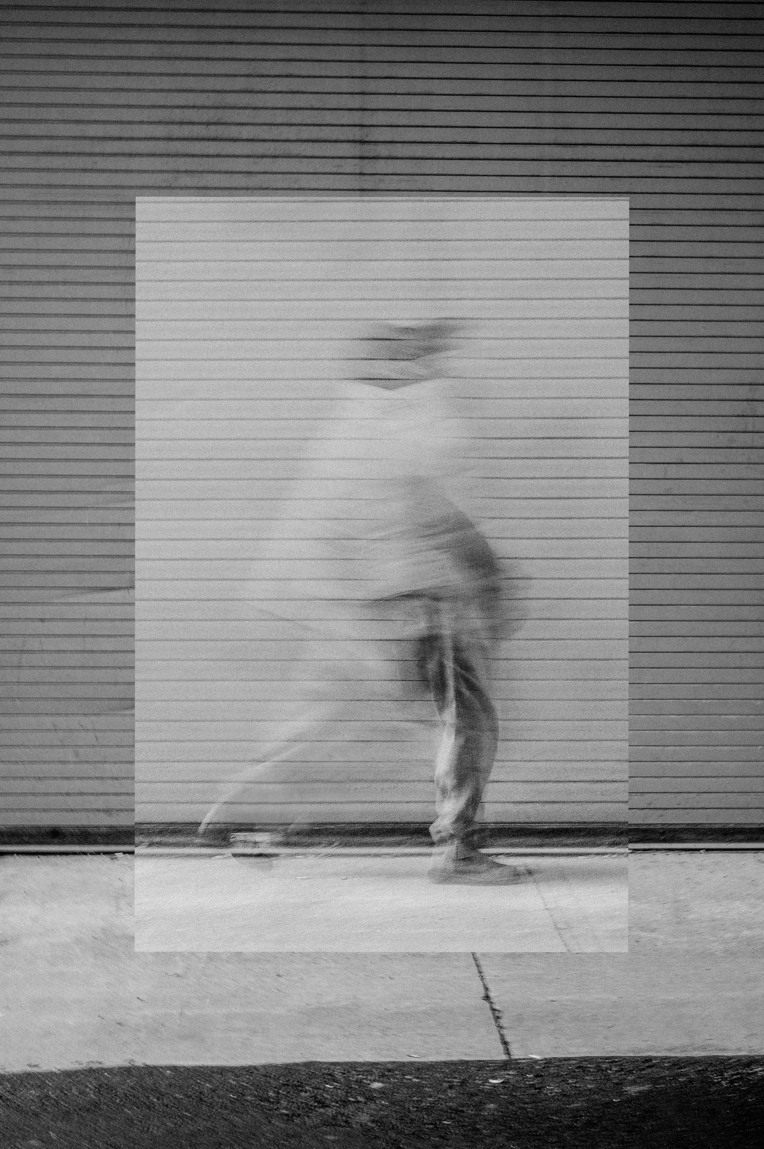 Blurred black and white image of a person skating on a sidewalk, in front of a metal roll-up door.