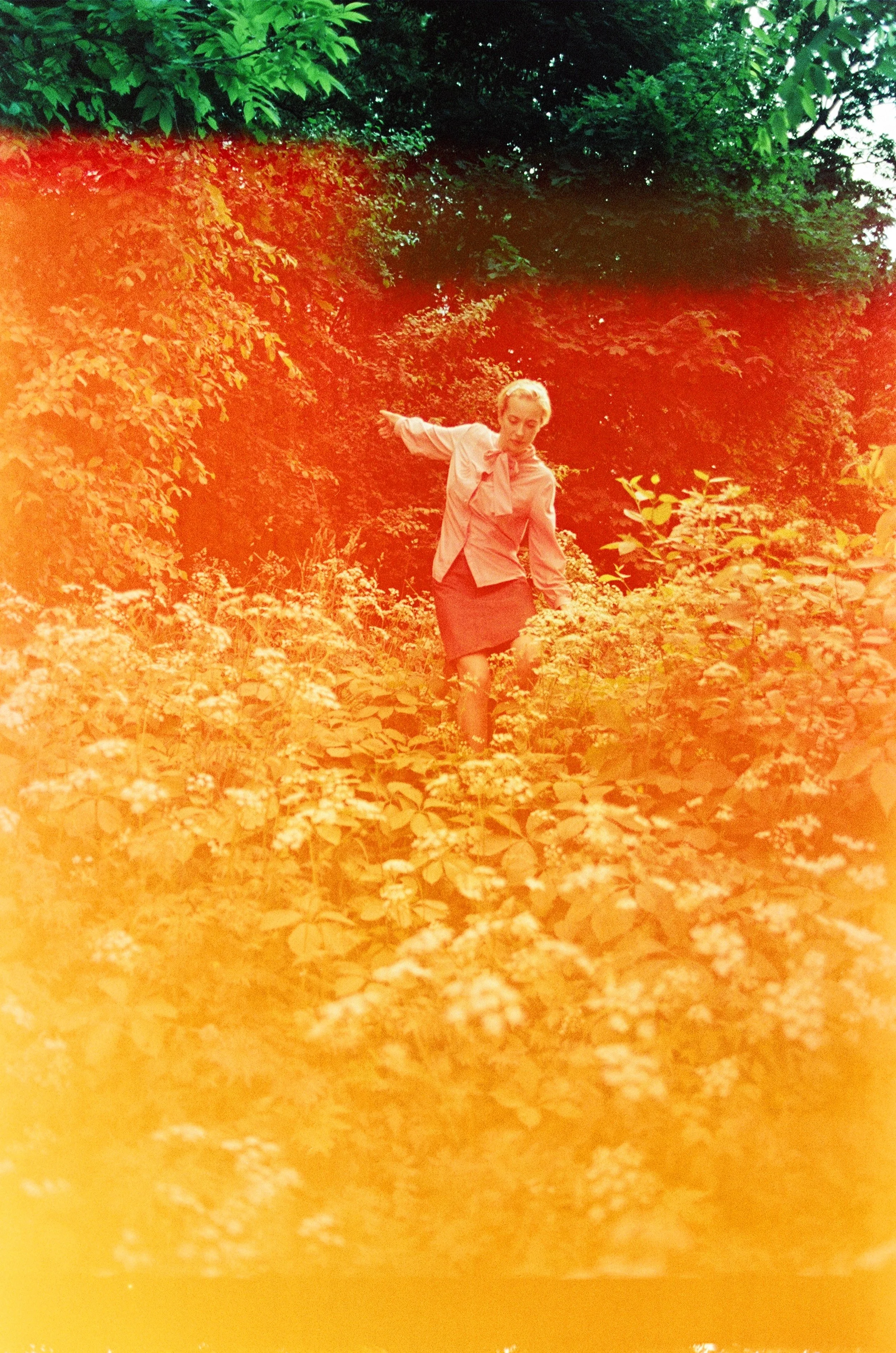 A woman in a pink blouse and dark skirt walking through a lush garden filled with white flowers, with green trees in the background.