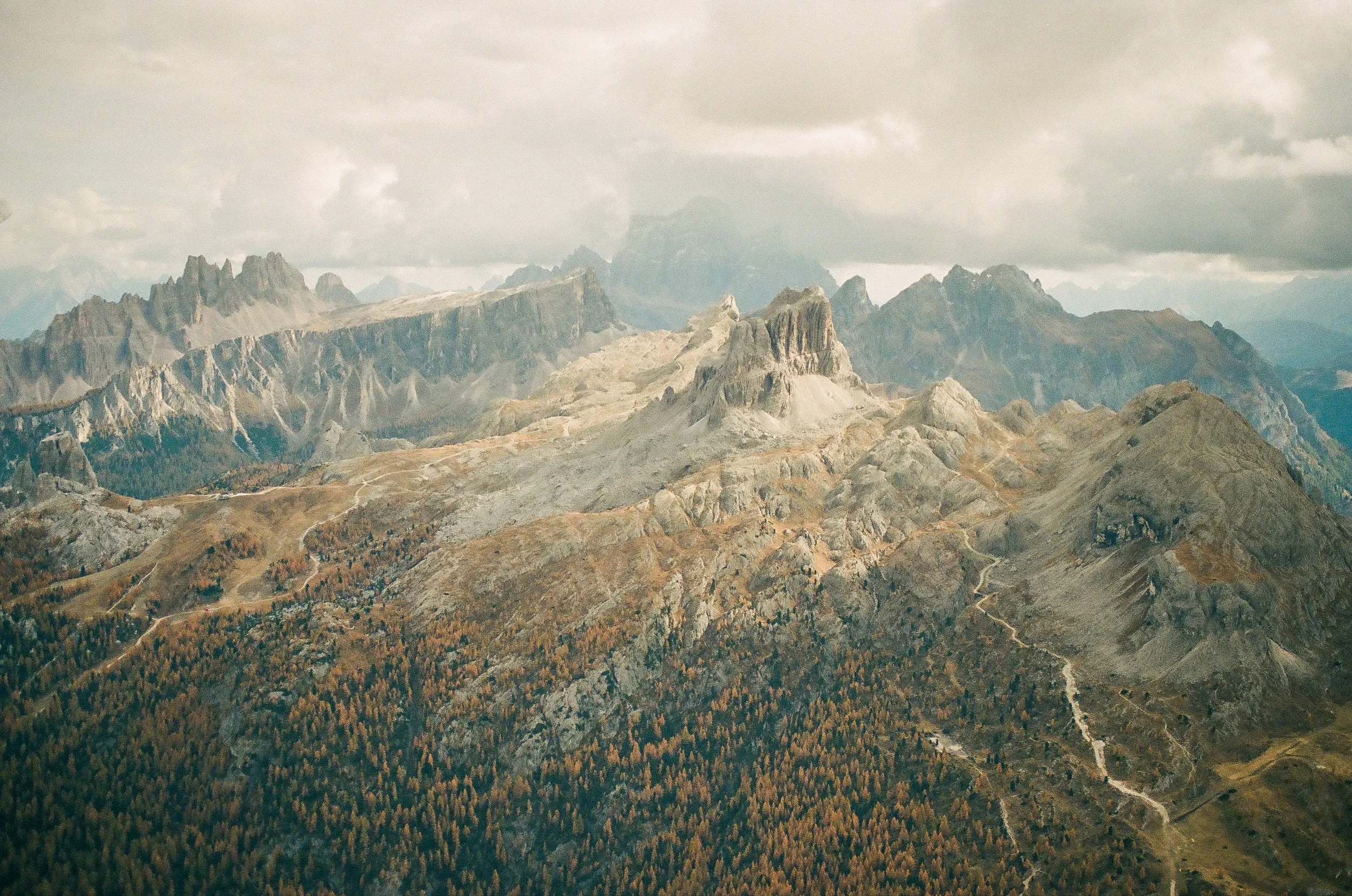Aerial view of rugged mountain landscape with rocky peaks, green forests, and overcast sky.