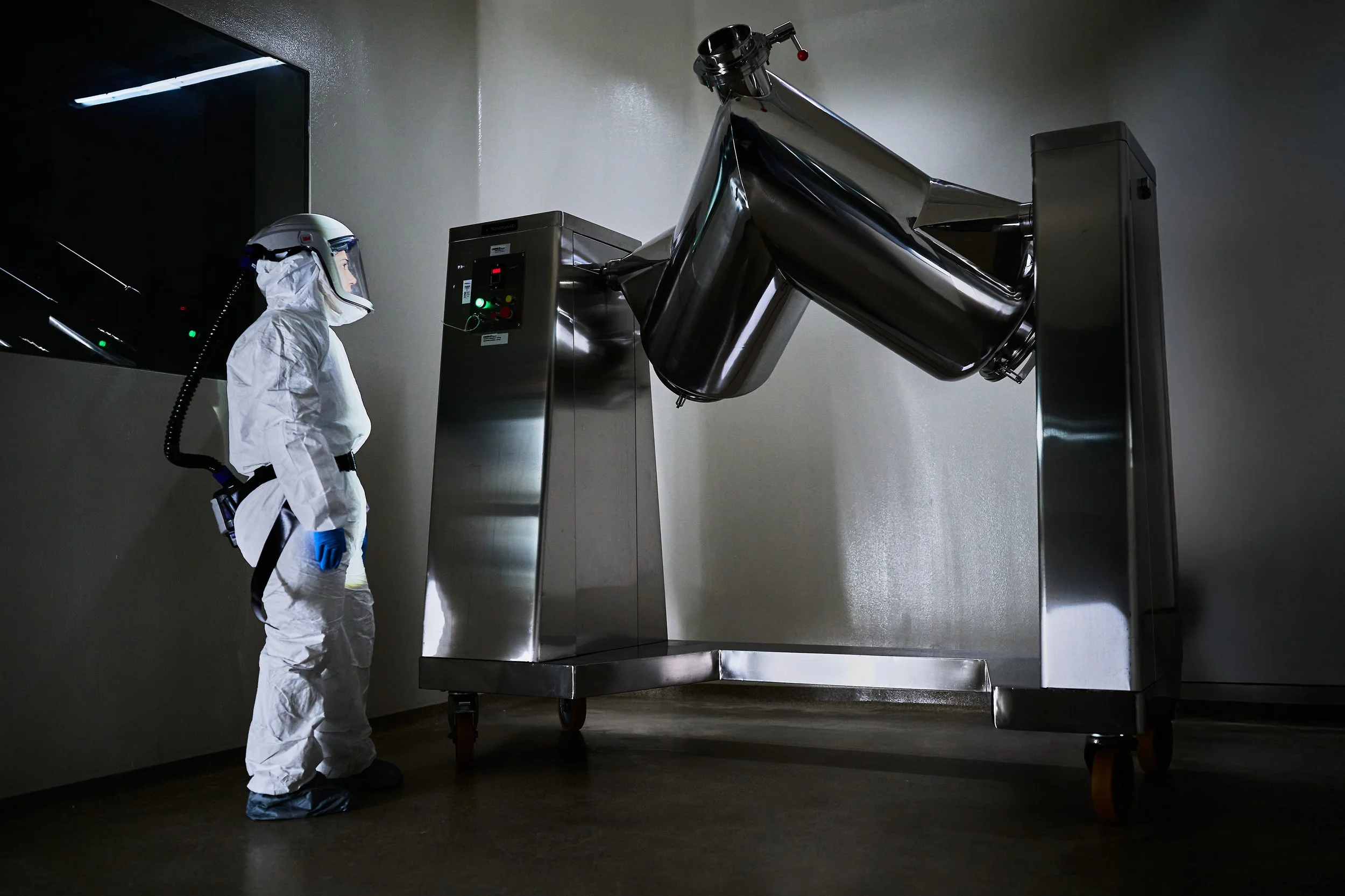A person in full protective gear, including a helmet, face shield, white coveralls, and blue gloves, standing next to a large stainless steel industrial machine in a dimly lit room.