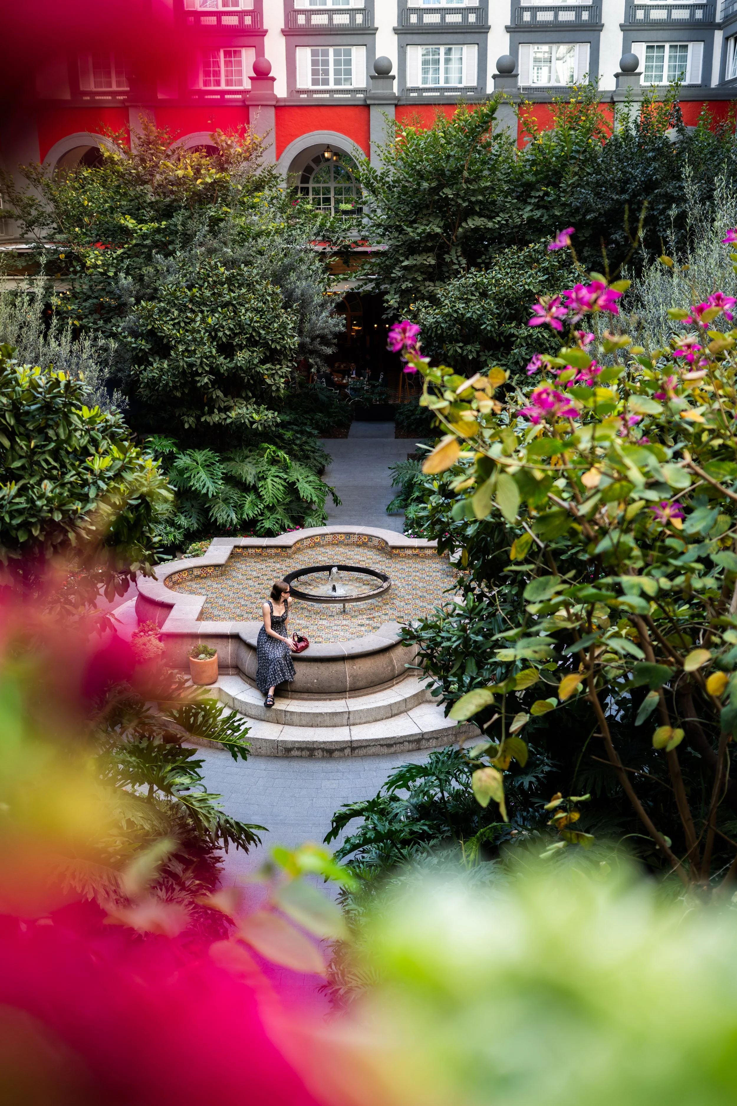 A woman in a black and white dress sits on the steps of a small fountain in a lush courtyard garden with pink and green foliage, in front of a multi-story building with red, grey, and white facade.