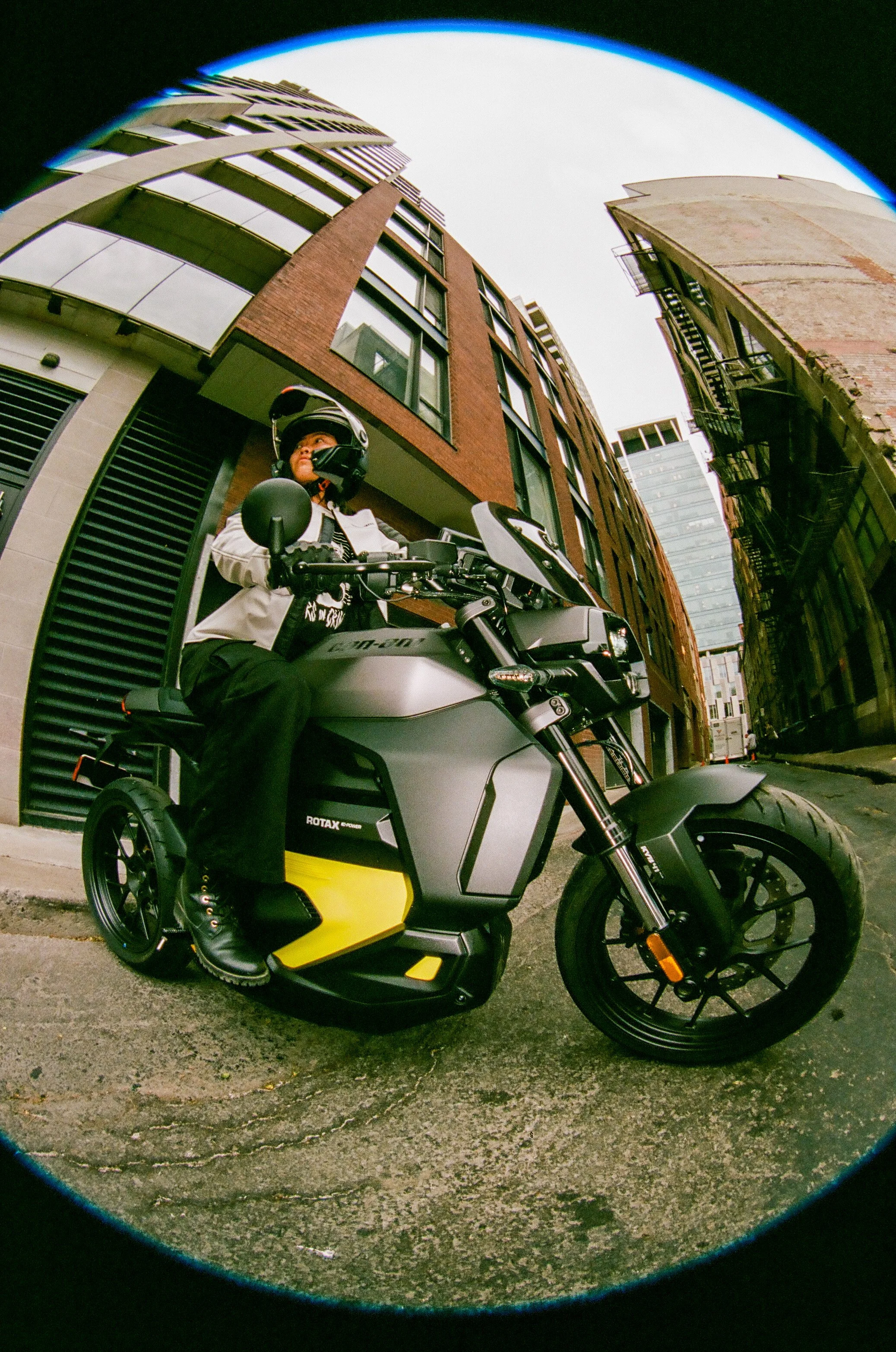 Person wearing a helmet and jacket sitting on an electric motorcycle on a city street with high-rise buildings and an overcast sky.