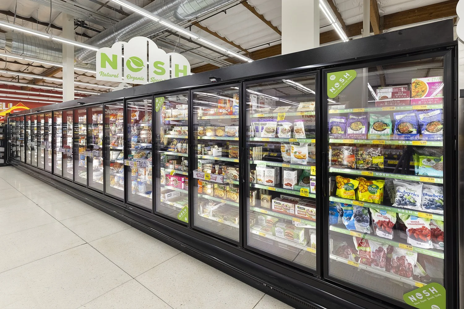Grocery store frozen food aisle with glass-door freezers containing frozen meals, vegetables, and berries, and a sign overhead reading 'Nosh'.