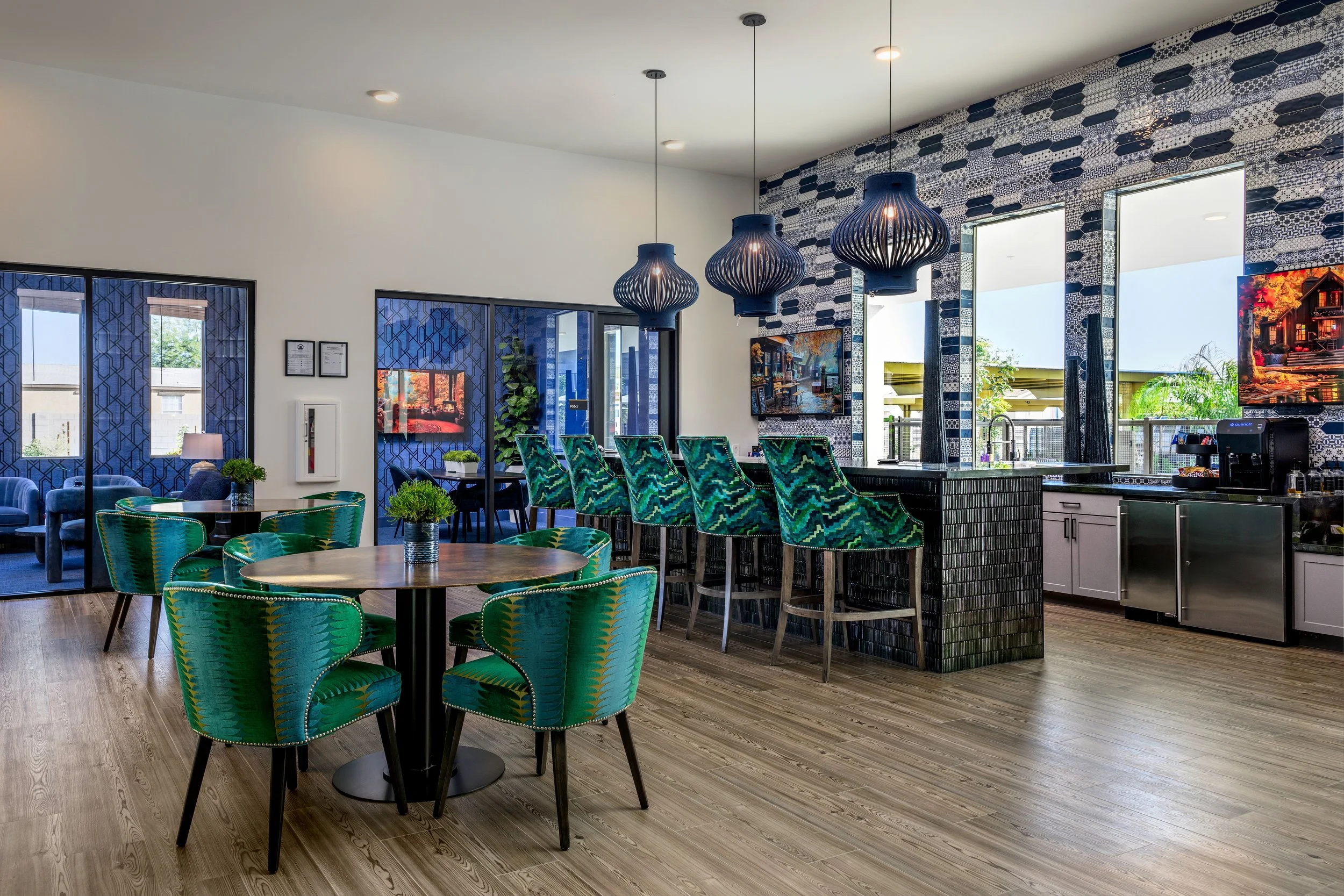 Interior of a modern bar and lounge area with colorful green chairs around a round table, a bar counter with green patterned barstools, decorative hanging pendant lights, large windows, and wall art.
