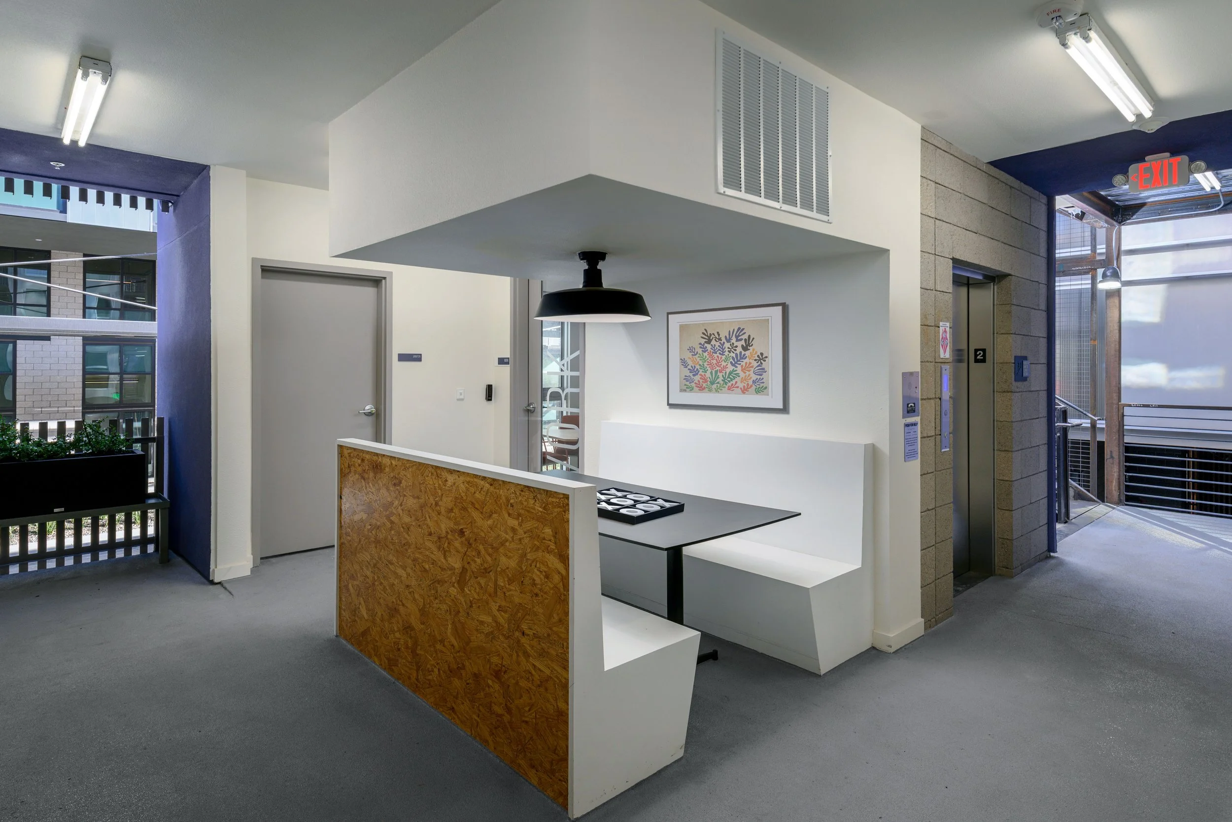 Interior of an office or common area featuring a seating nook with white bench and table, decorated with wall art, adjacent to elevator and exit door, with bright lighting and modern decor.