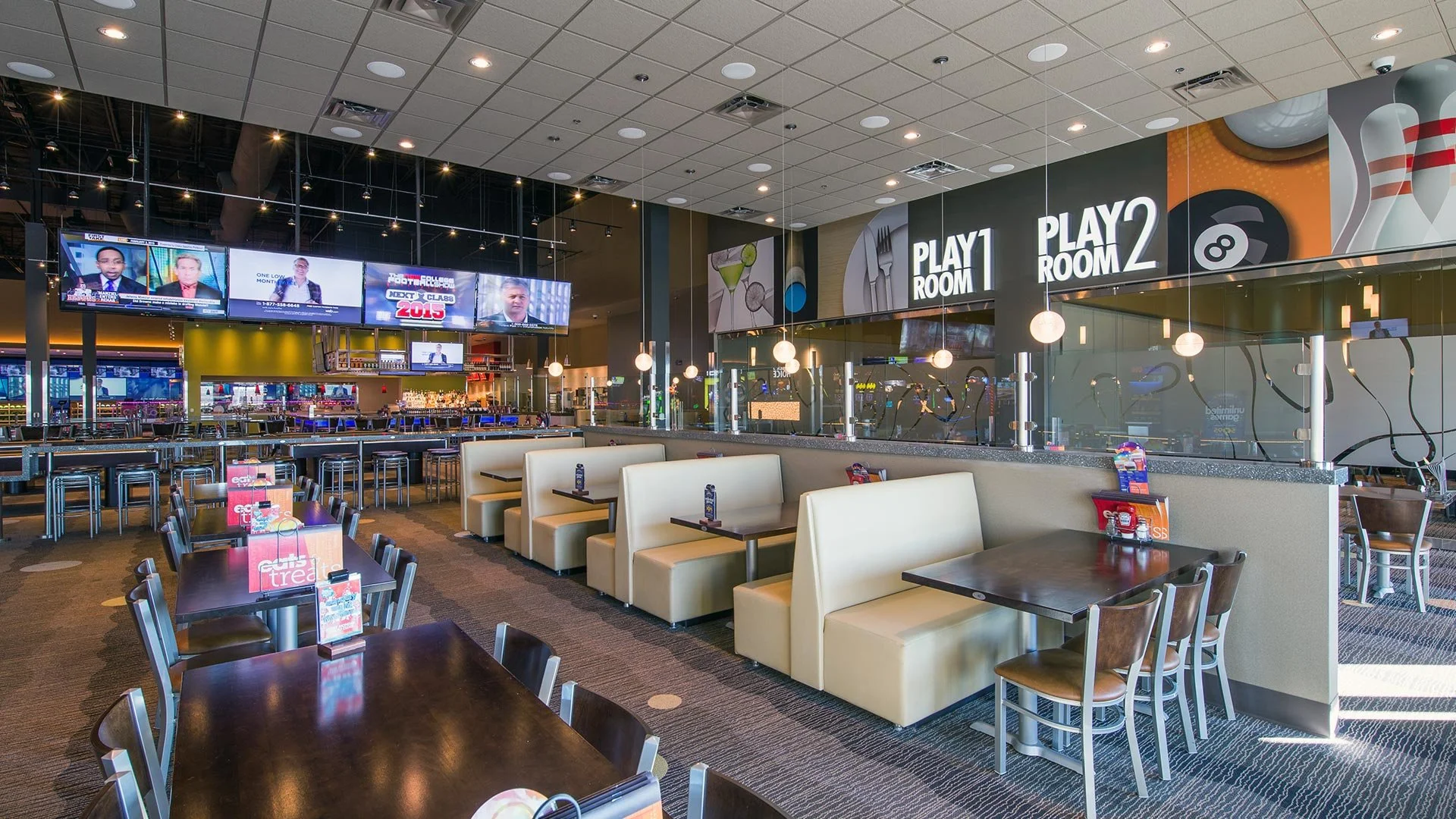 Interior of a modern bowling alley with multiple seating areas, sports screens, and a snack bar in the background.