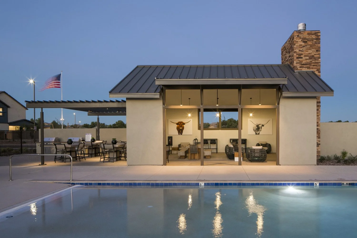 Poolside building with outdoor seating, mounted bull and cow skull art, illuminated interior, brick chimney, American flag on a pole, evening sky.