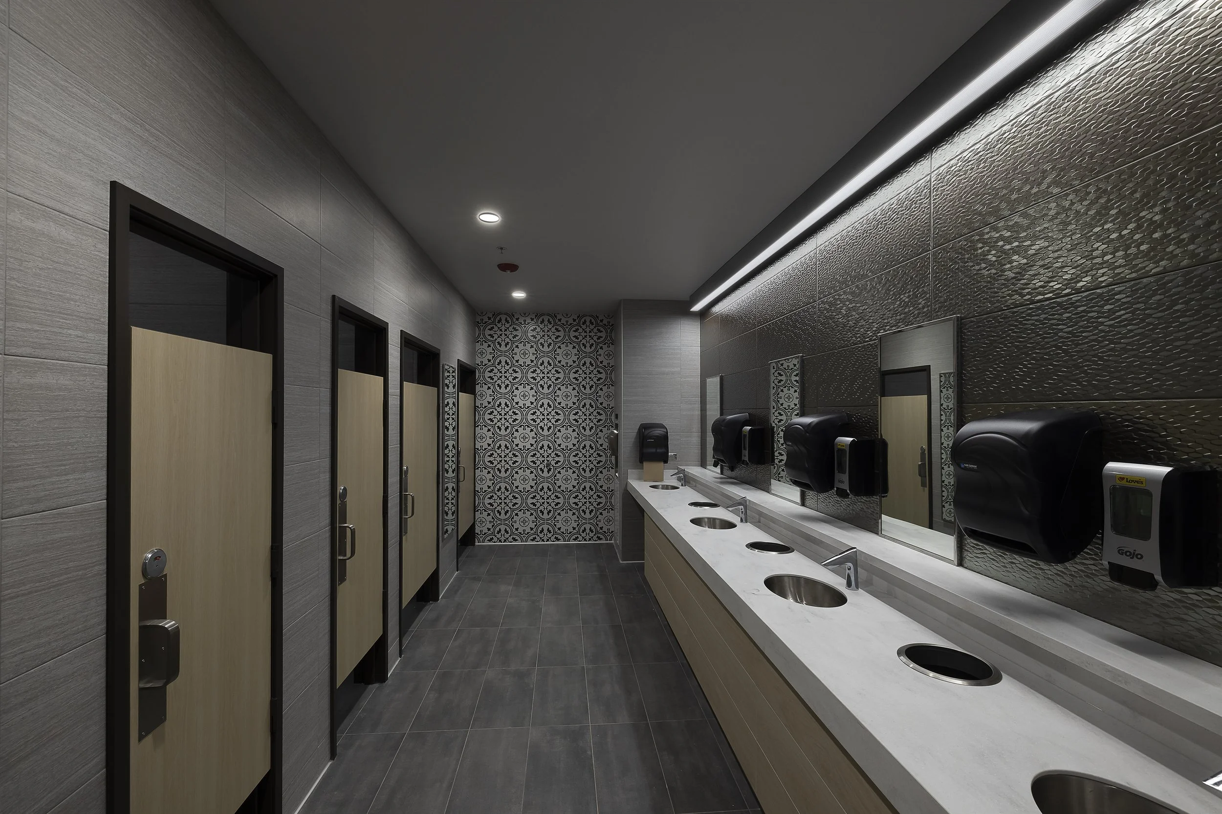 Public restroom with multiple toilet stalls on the left and a row of sinks with soap dispensers and mirrors on the right, decorated with gray tiles and patterned wall tiles at the far end.