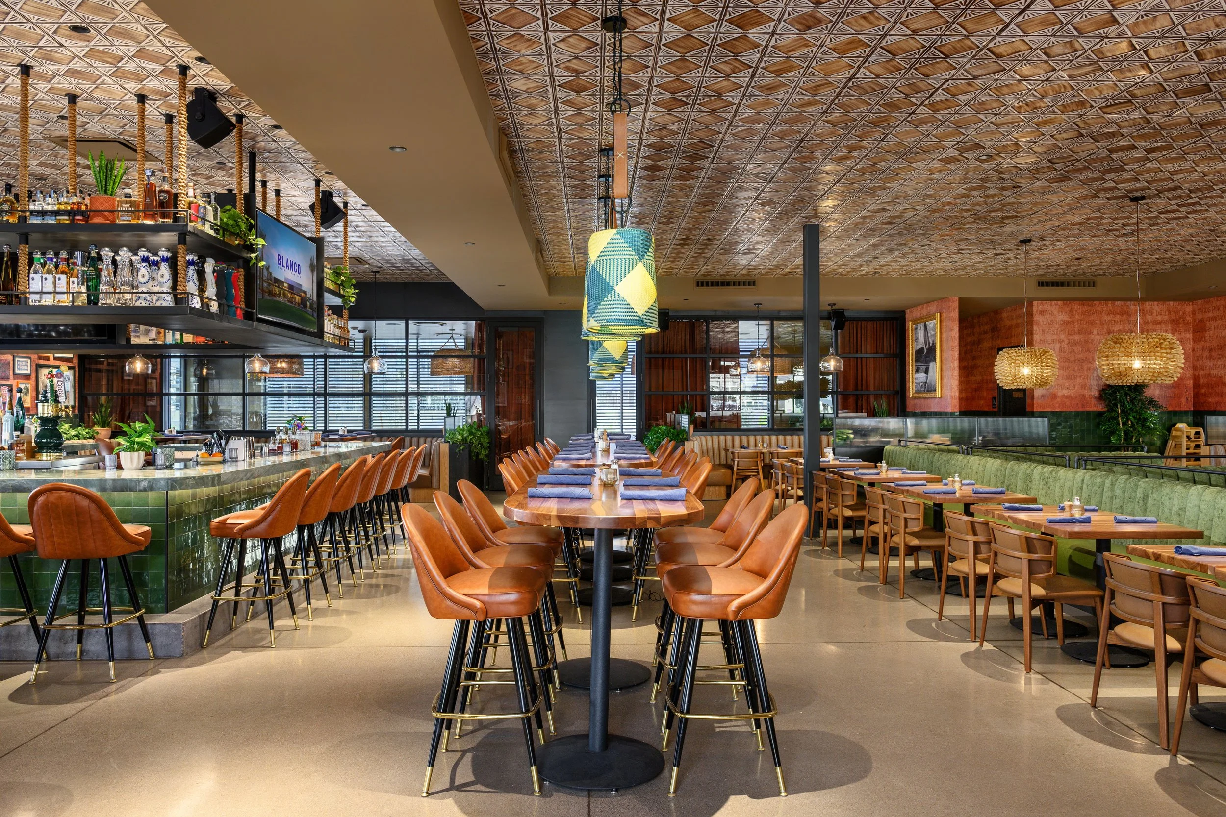 Interior of a restaurant with a bar on the left, high chairs at the bar counter, and a long table in the center with brown leather chairs. Seating on the right includes a green cushioned banquette and wooden chairs with tables set with blue napkins. 