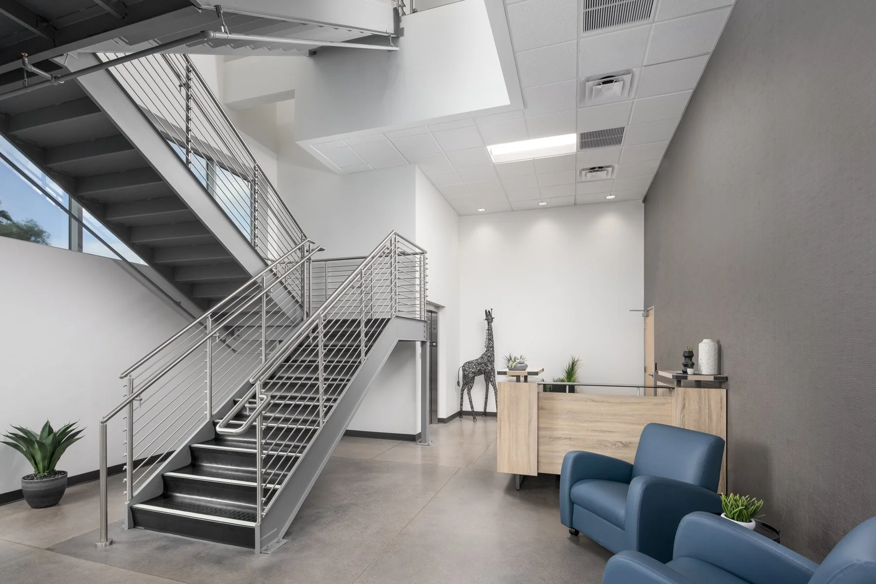 Modern office lobby featuring a metal staircase, a blue armchair, a wooden reception desk, and decorative plants.