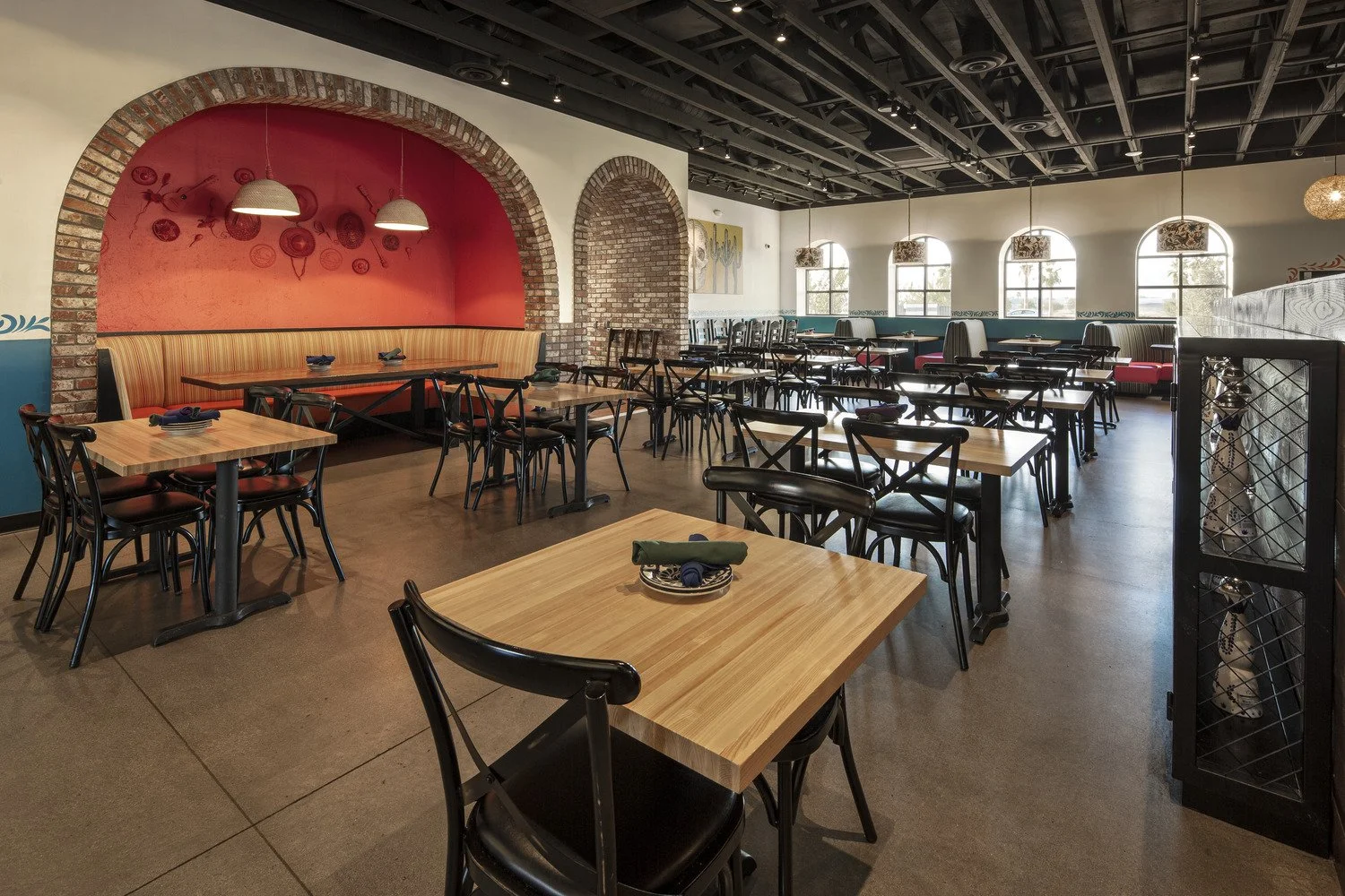 Empty restaurant dining area with wooden tables, black chairs, brick accents, and large arched windows.